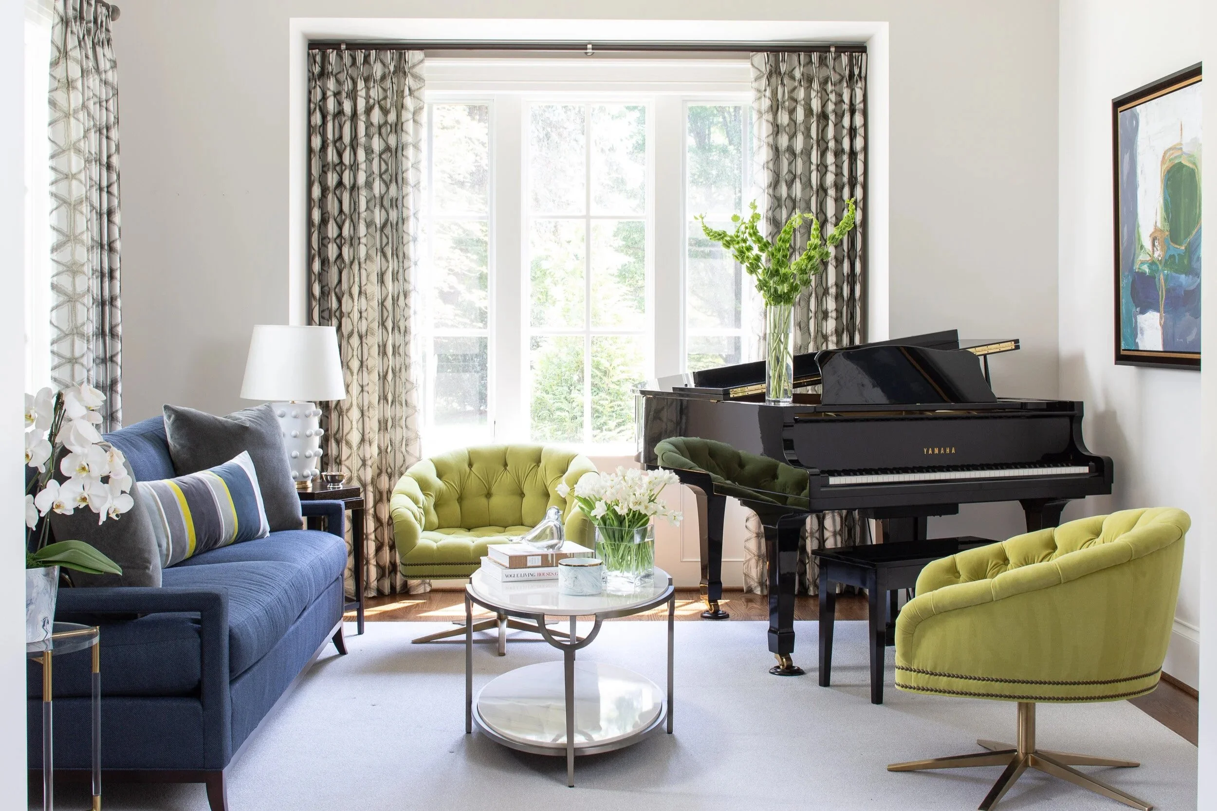 Living room with a blue sofa, gray and striped cushions, a white side table with a lamp, a round coffee table with books, a vase with white flowers, a black grand piano with a glass vase on top, and a yellow velvet chair, all positioned in front of large windows with patterned curtains.