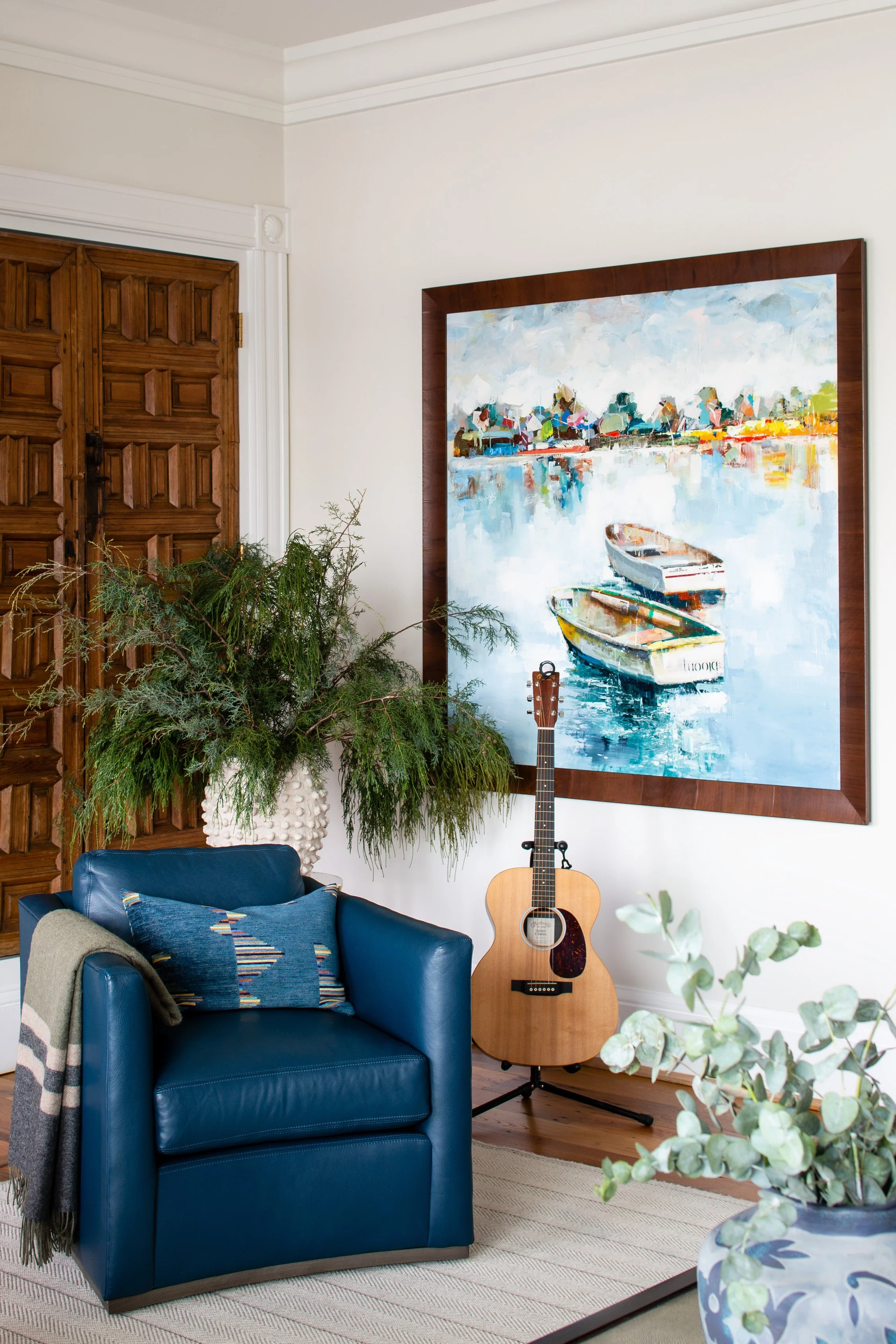 Interior of a living room with a blue armchair, a pillow, a blanket, a large abstract painting of boats on water, a wooden acoustic guitar, a large potted plant, and a decorative ceramic pot with eucalyptus.