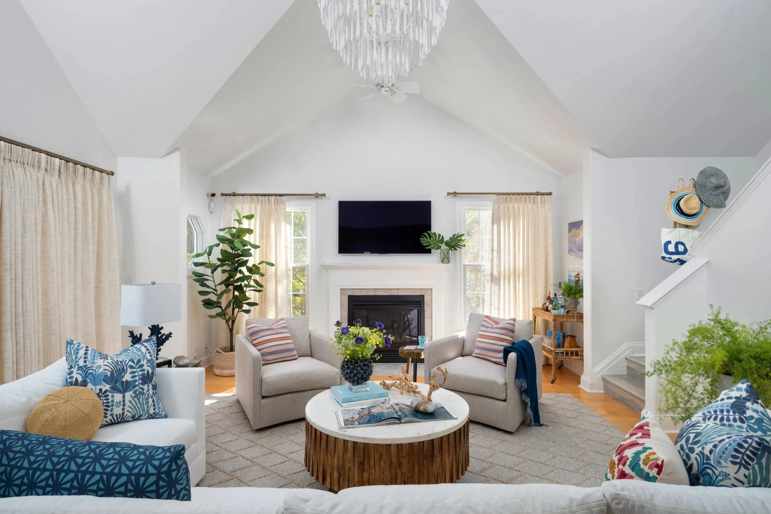 Bright living room with white walls, high vaulted ceiling, and large windows with beige curtains. Contains a fireplace with a mounted TV above, surrounded by two armchairs with striped pillows, a white sofa with blue pillows, and a round coffee table with books, a flower vase, and decorative objects. Green plants are on either side, and a chandelier hangs from the ceiling.