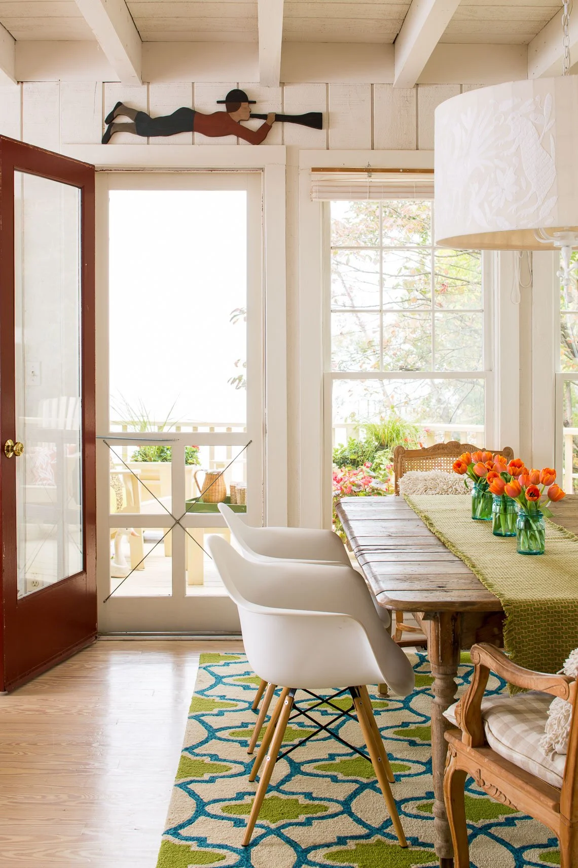 Bright dining room with wooden table, white chairs, orange tulip flowers in green vases, and large windows with sunlight.