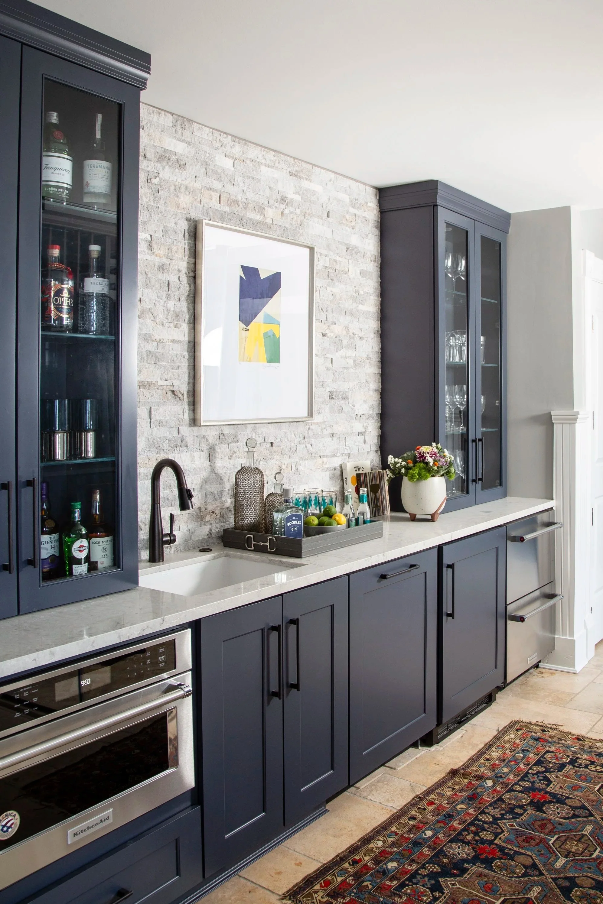 Modern kitchen with dark blue cabinets, light stone countertop, white brick accent wall, and decorative items including bottles, plants, and artwork.