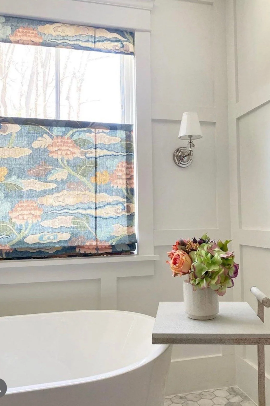 A bright bathroom with a white freestanding bathtub, a window with patterned fabric curtains, a small white table with a flower vase, and a wall-mounted lamp.