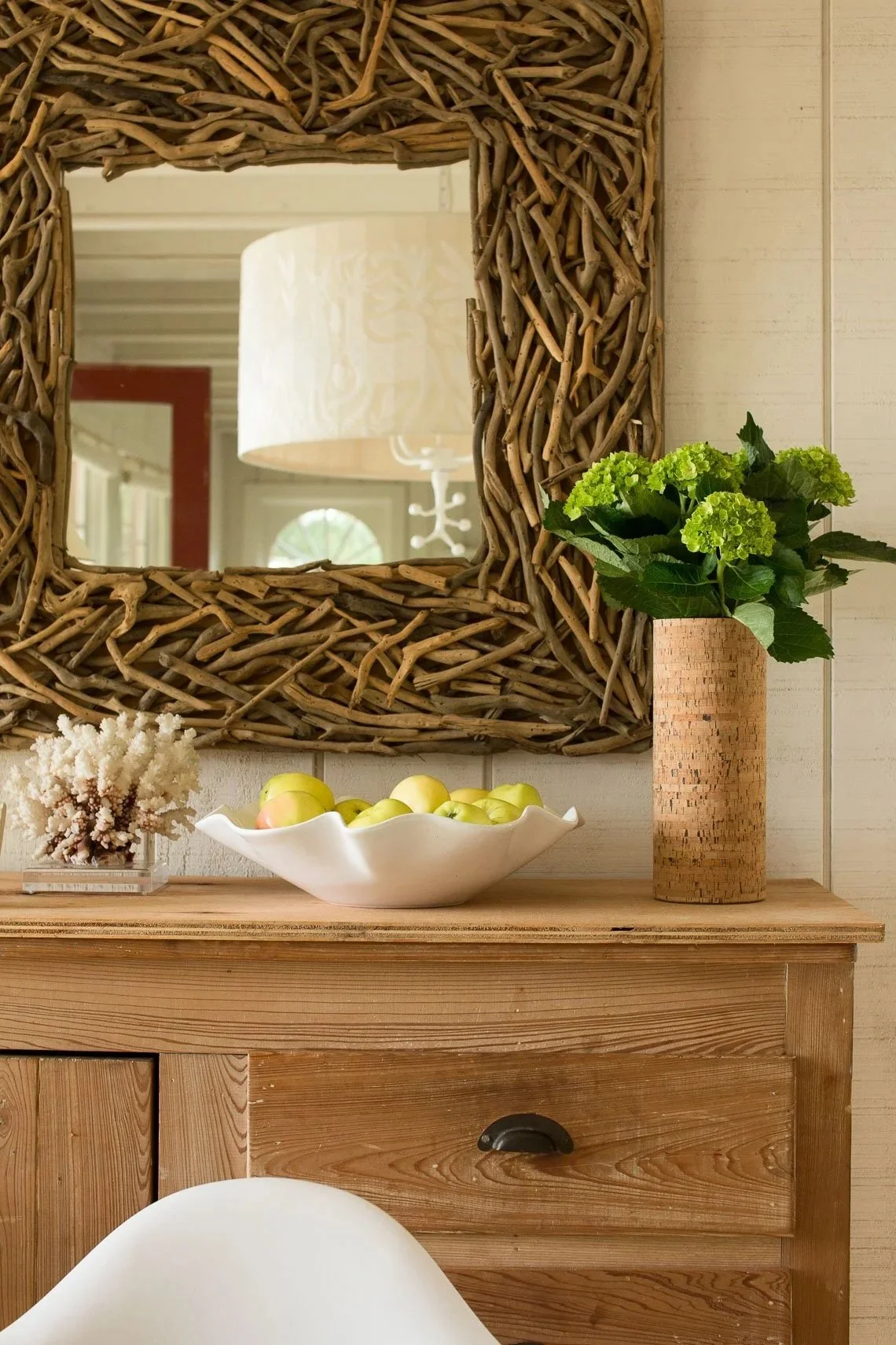 Decorative wooden console table with a large wicker-framed mirror, a green hydrangea plant in a cork vase, a white shell-shaped bowl with yellow apples, and a small coral branch in a glass container, in a rustic farmhouse-style interior.