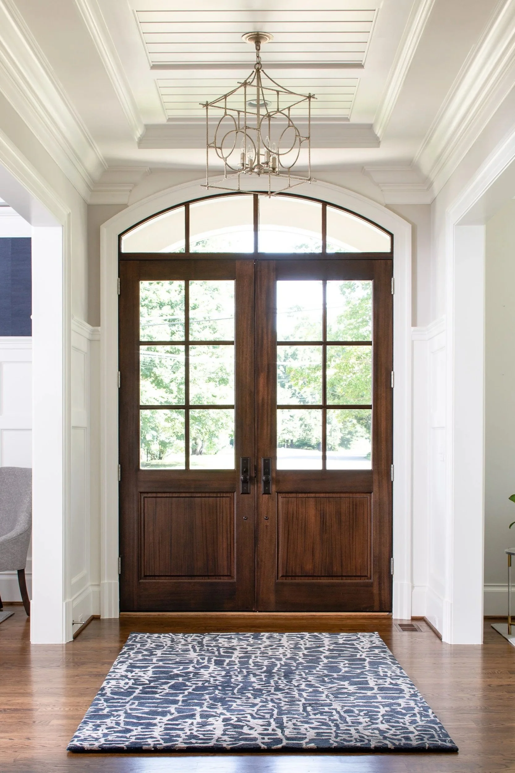 Front door with glass panels, wooden frame, arched window above, and a patterned rug on hardwood floor in entryway.