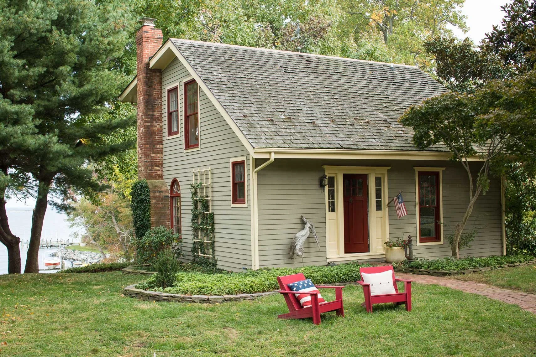 A house with gray siding, red-trimmed windows, and a brick chimney, surrounded by trees and a lawn with two red Adirondack chairs, one with an American flag pillow.