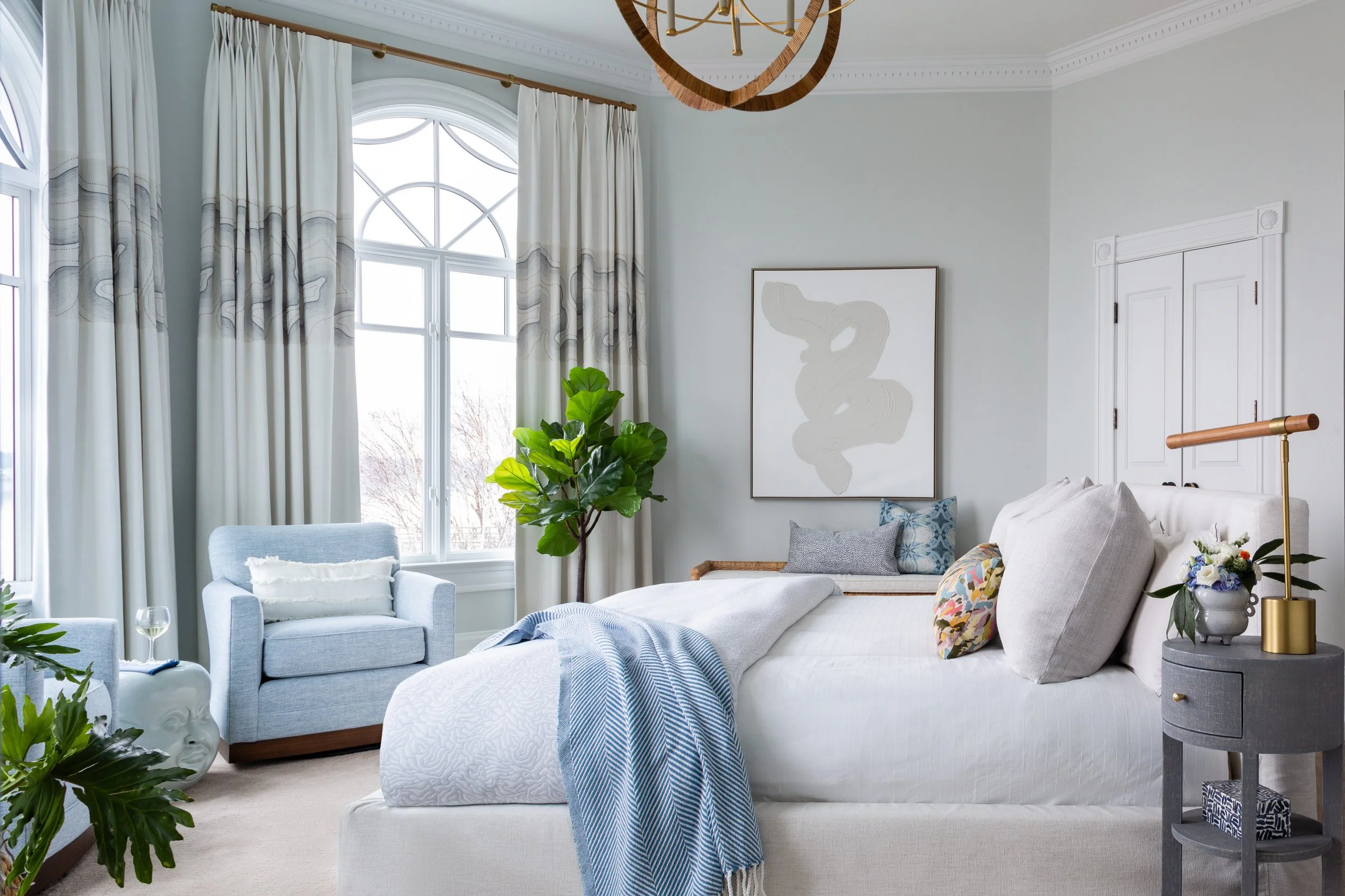 Bright bedroom with large arched windows, white curtains, light-colored walls, a bed with multiple pillows and a blue throw, a gray side table with a flower vase and lamp, a blue armchair with a pillow, and green indoor plants.