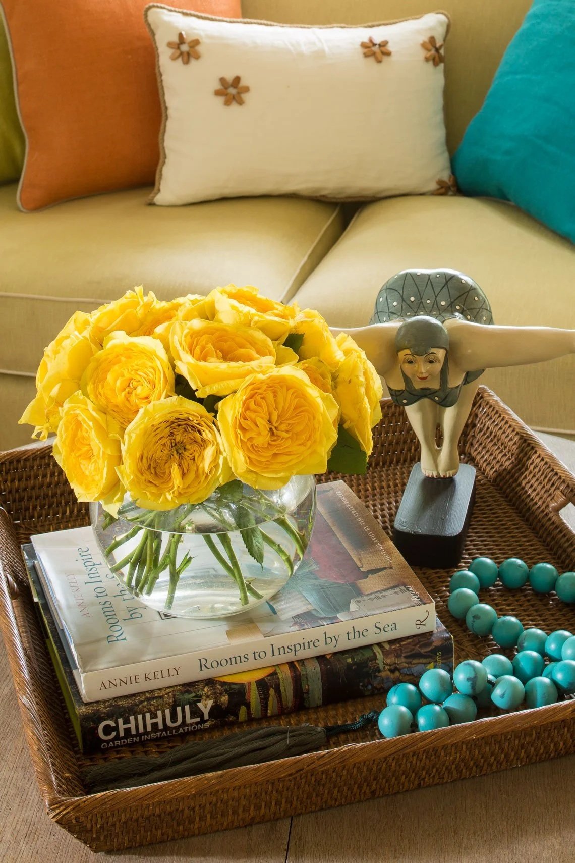 A wicker tray on a table holding a clear glass vase with yellow roses, a statue of a woman with arms outstretched, a stack of books including "Rooms to Inspire by the Sea," and a turquoise bead necklace. In the background, a beige sofa with colorful pillows.