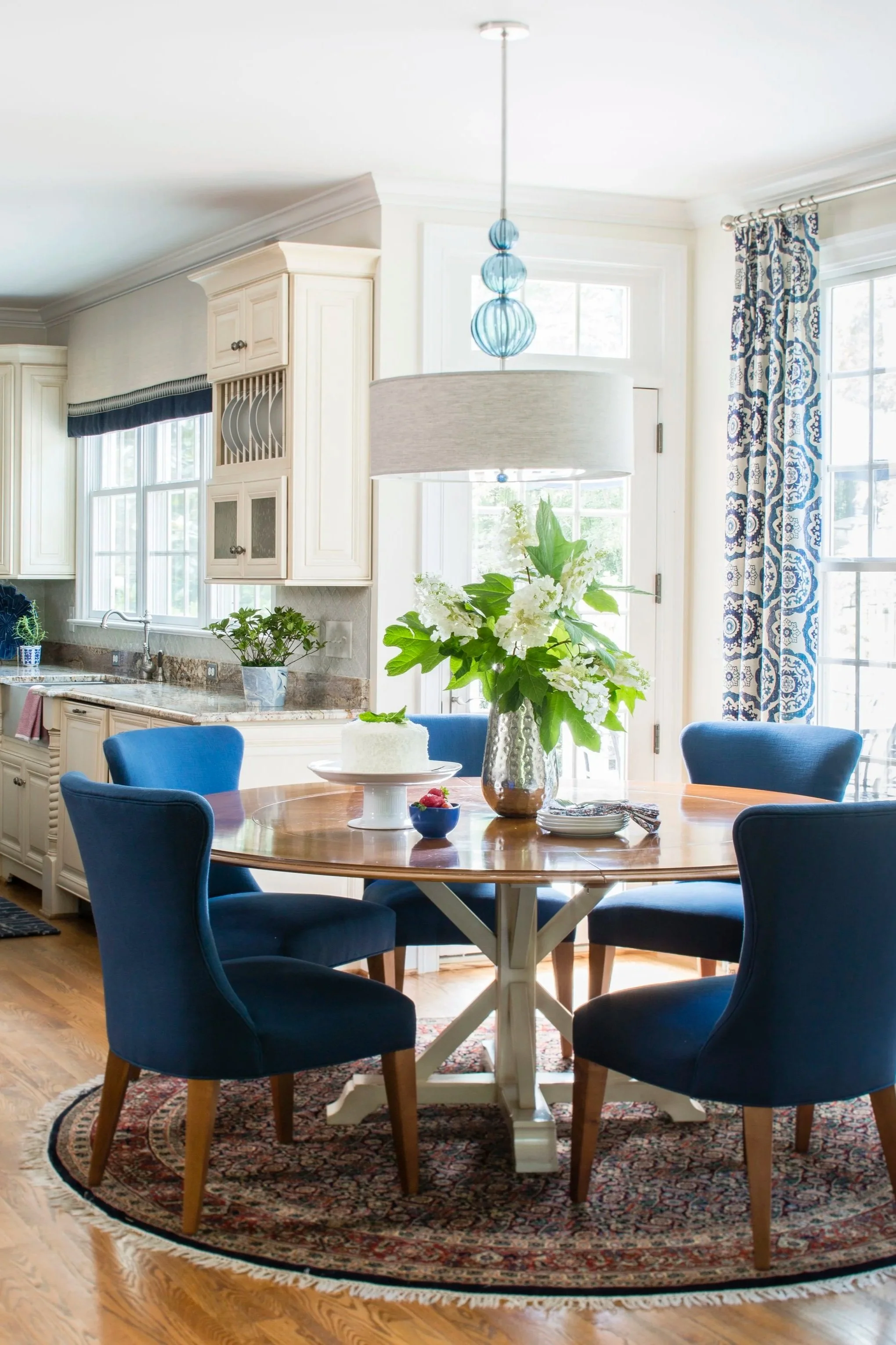 A bright, cozy kitchen and dining area with a round wooden table, blue upholstered chairs, a white vase with large white flowers, a cake on a stand, and a small bowl of berries. The space features large windows with patterned curtains, white cabinetry, and wooden flooring.