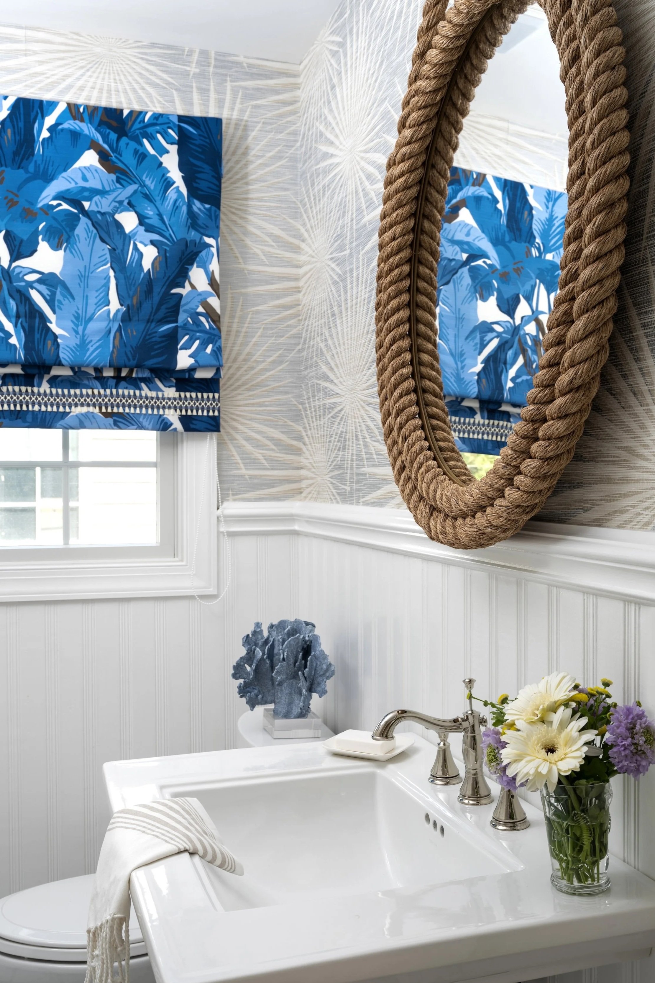 Bathroom with blue tropical patterned window valance, textured mirror with rope frame, white wainscoting, soap, flowers, and blue coral sculpture on a small table.