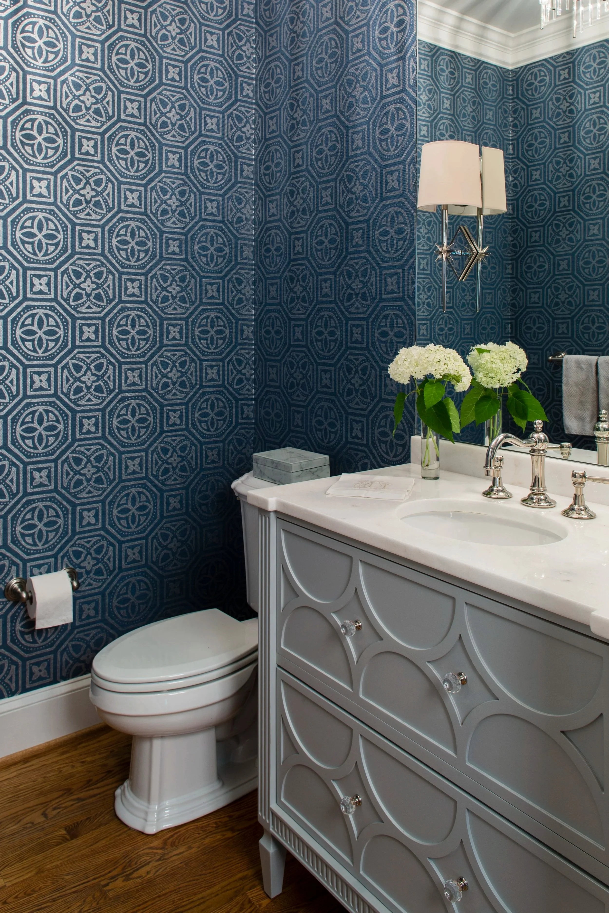 A bathroom with a blue patterned wallpaper, a white vanity with a marble top, a mirror, a vase with white hydrangeas, a wall-mounted light, and a toilet with a chrome toilet paper holder.