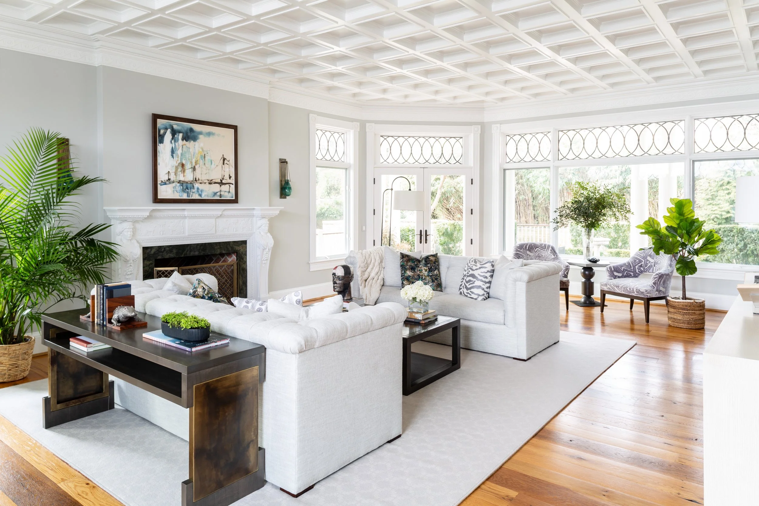 Bright living room with white sofas, potted plants, large windows, hardwood floors, and decorative fireplace with artwork above.