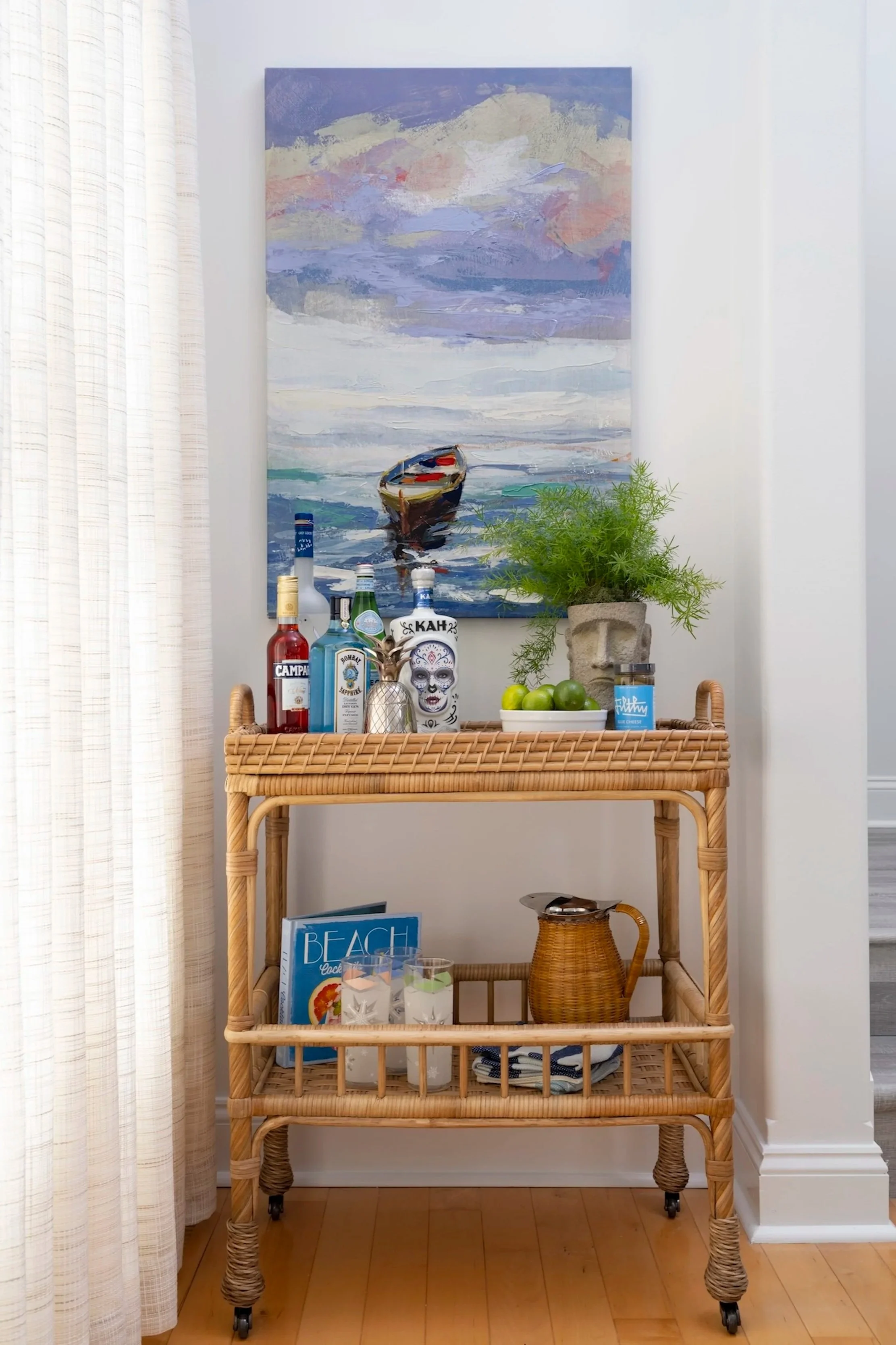 A wicker bar cart with a glass top, holding liquor bottles, a small pineapple sculpture, a potted plant, and a bowl of green apples. Below, there are cocktail glasses with drinks, a woven pitcher, and a beach magazine. A colorful abstract painting of a boat and ocean is hung on the wall above, with white curtains on the left.