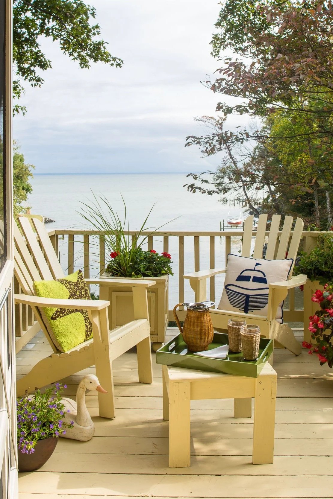 A cozy outdoor porch overlooking a calm body of water with trees and cloudy sky in the background. The porch has two white wooden chairs with colorful cushions, a small table with decorative items, and potted plants, creating a relaxing lakeside setting.