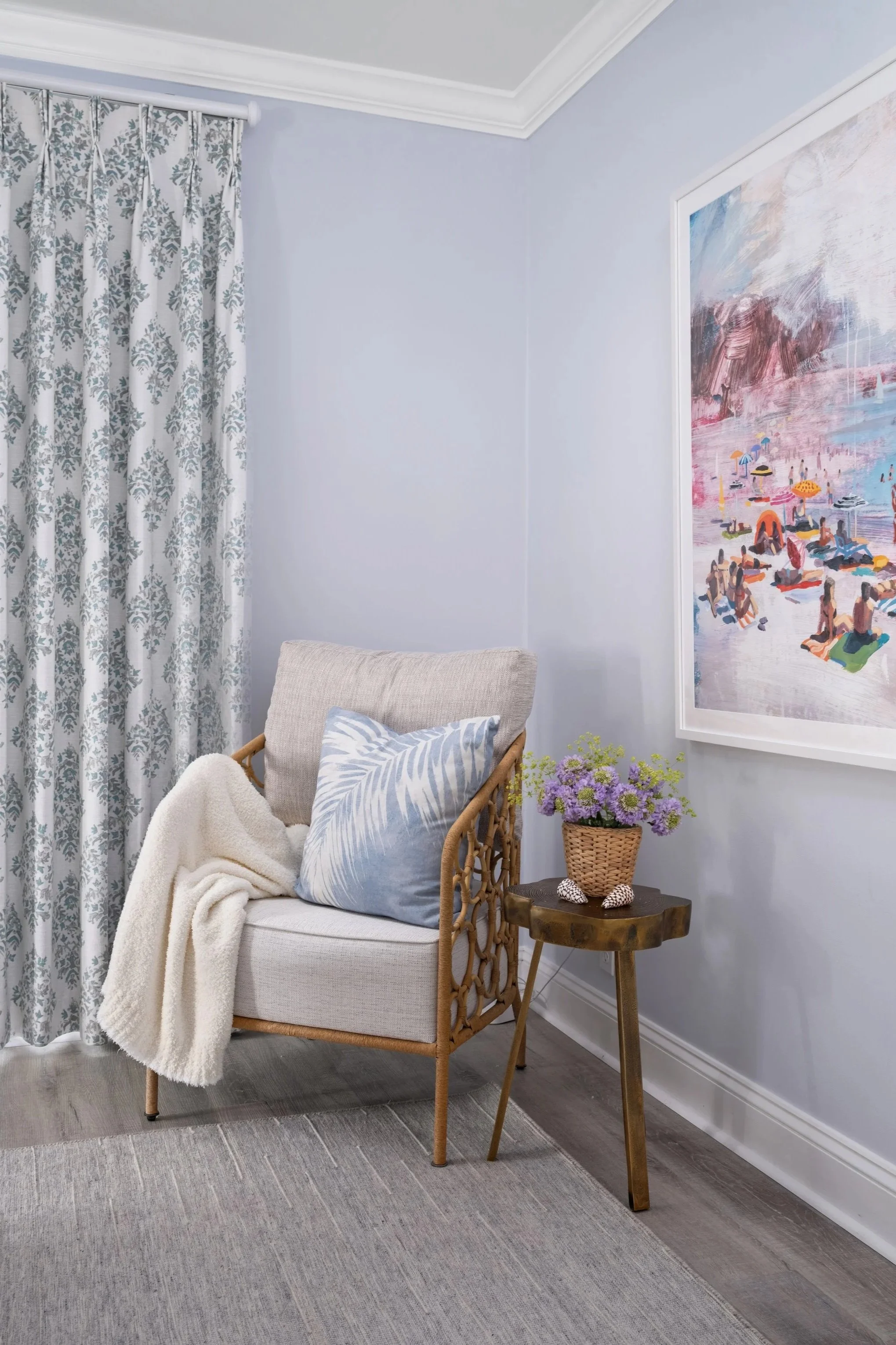 A cozy corner with a rattan armchair featuring a gray cushion, a pillow with a blue and white leaf pattern, a white blanket, and a small wooden side table with a woven basket of purple flowers, beside a large colorful beach scene painting and a window with gray floral curtains.