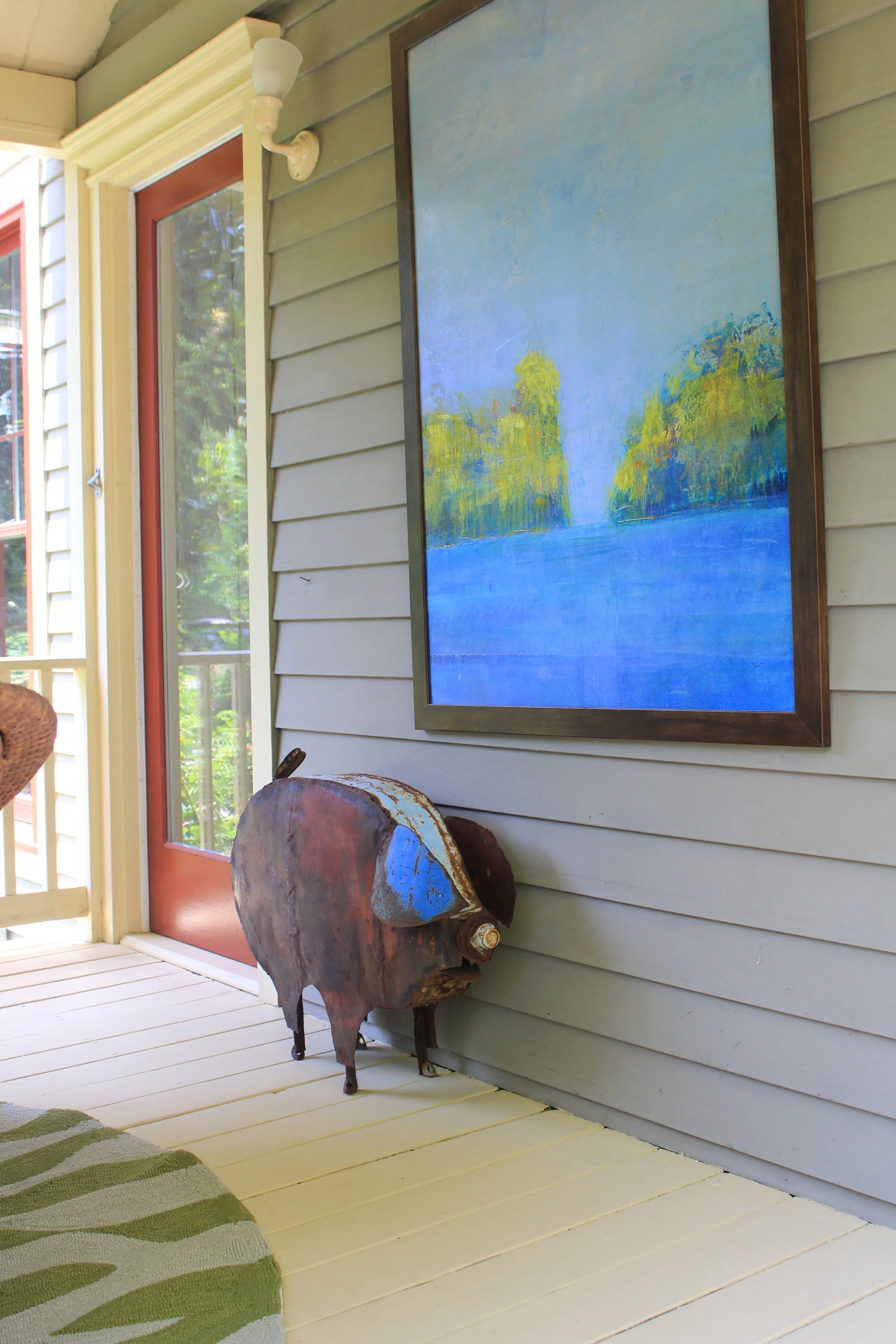 A porch or entryway with a green wooden diamond-plank floor, beige body siding, and a glass door with a red frame. There is a painting of a river scene with blue water and green trees on the wall, and a rustic pig-shaped metal sculpture near the wall.