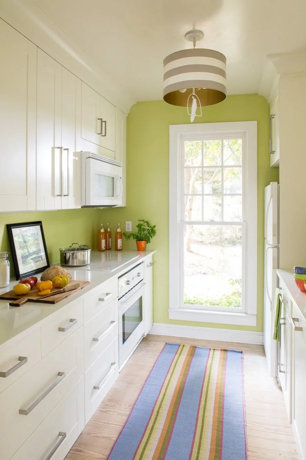 Bright kitchen with white cabinets, a green painted wall, and a large window letting in natural light. There is a striped rug on the wooden floor and various kitchen items on the countertops.
