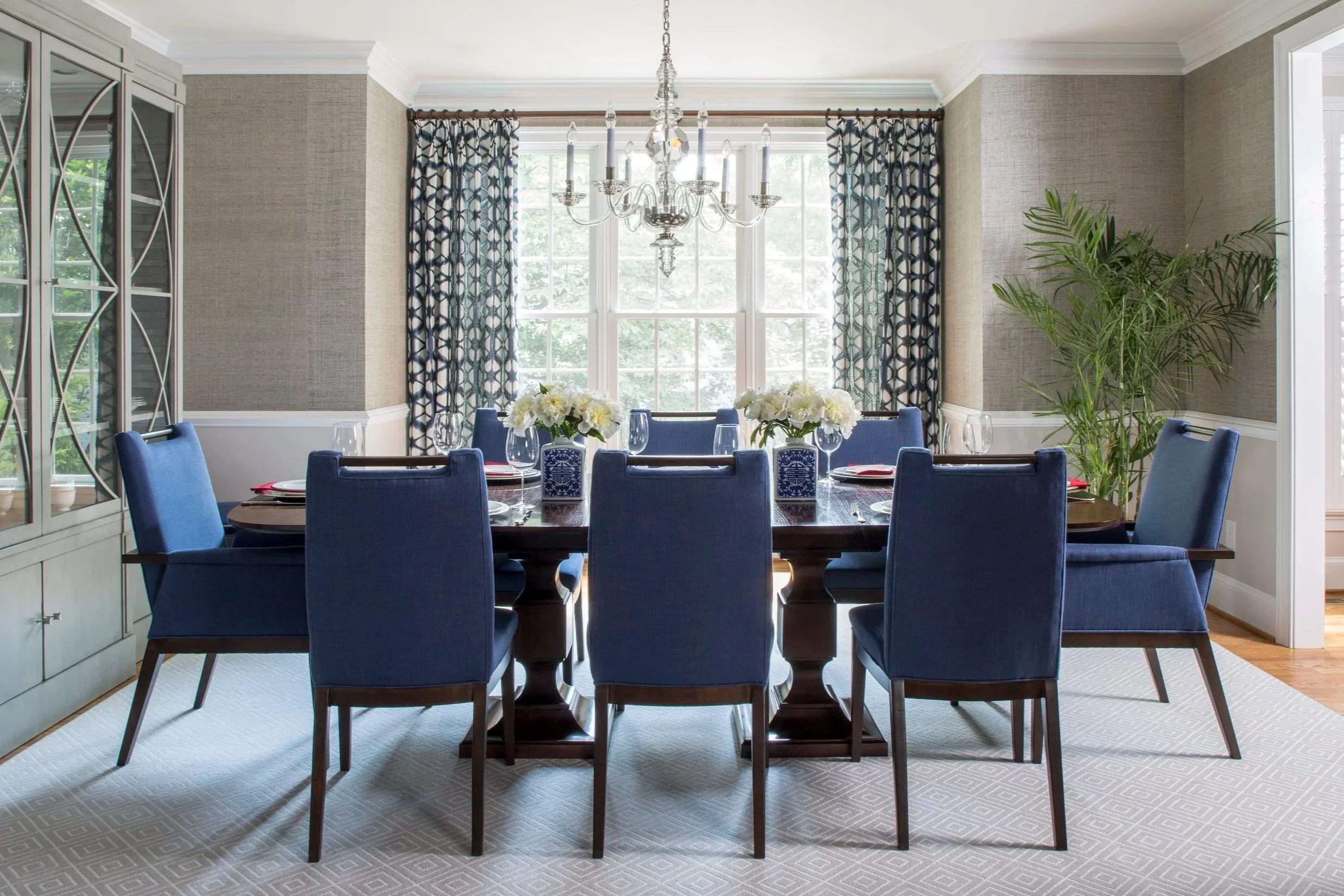 Dining room with a wooden table set for six, blue upholstered chairs, white floral centerpieces, large window with patterned curtains, chandelier hanging above, built-in cabinet on the left, and a large green plant on the right.