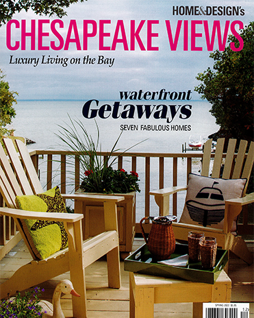 A furnished deck overlooking a bay with two wooden chairs, a side table, and various potted plants with a view of water and trees in the background.
