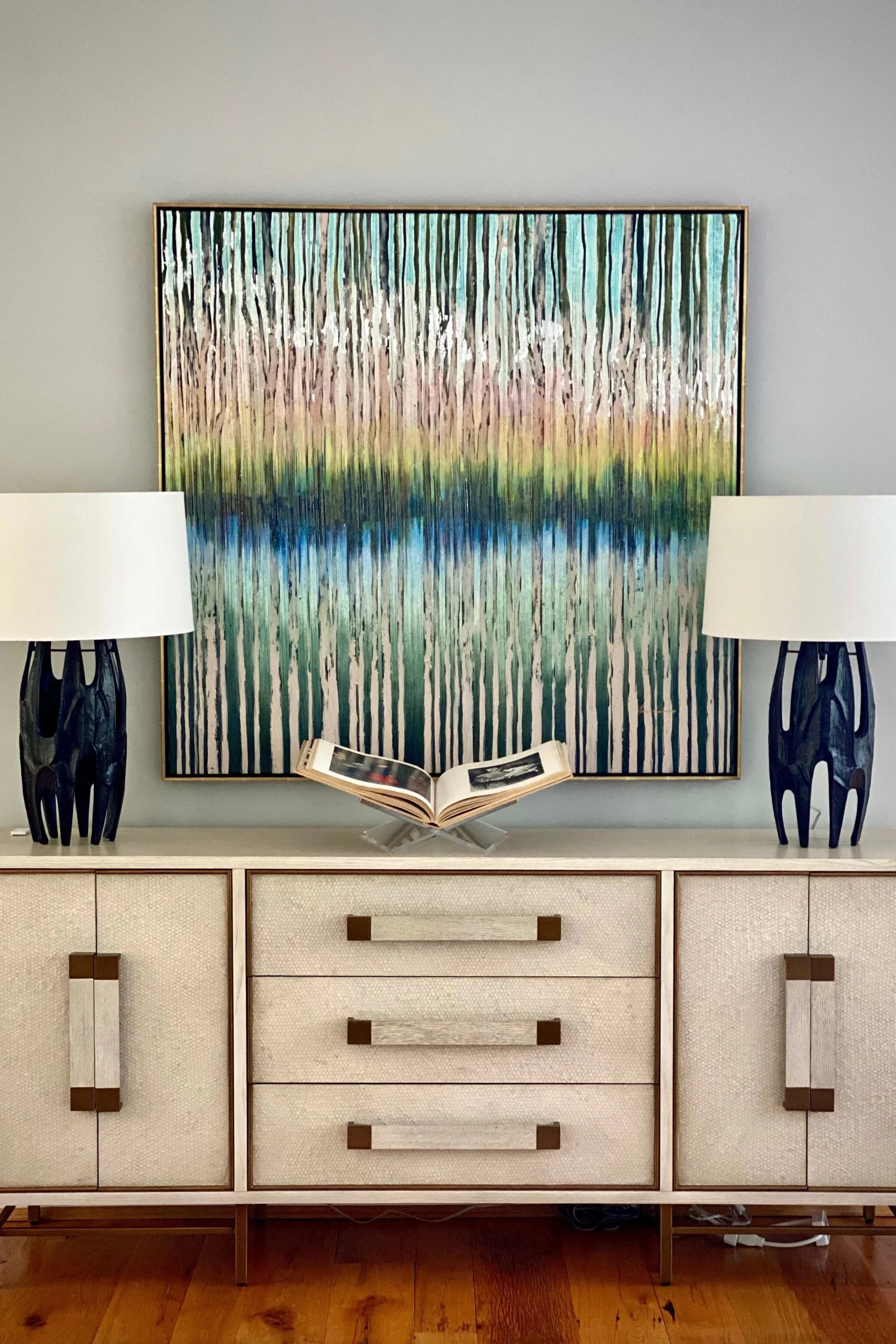 A modern credenza with two table lamps on either side, an open book in the middle, a colorful abstract painting behind, all set against a light gray wall and hardwood floor.