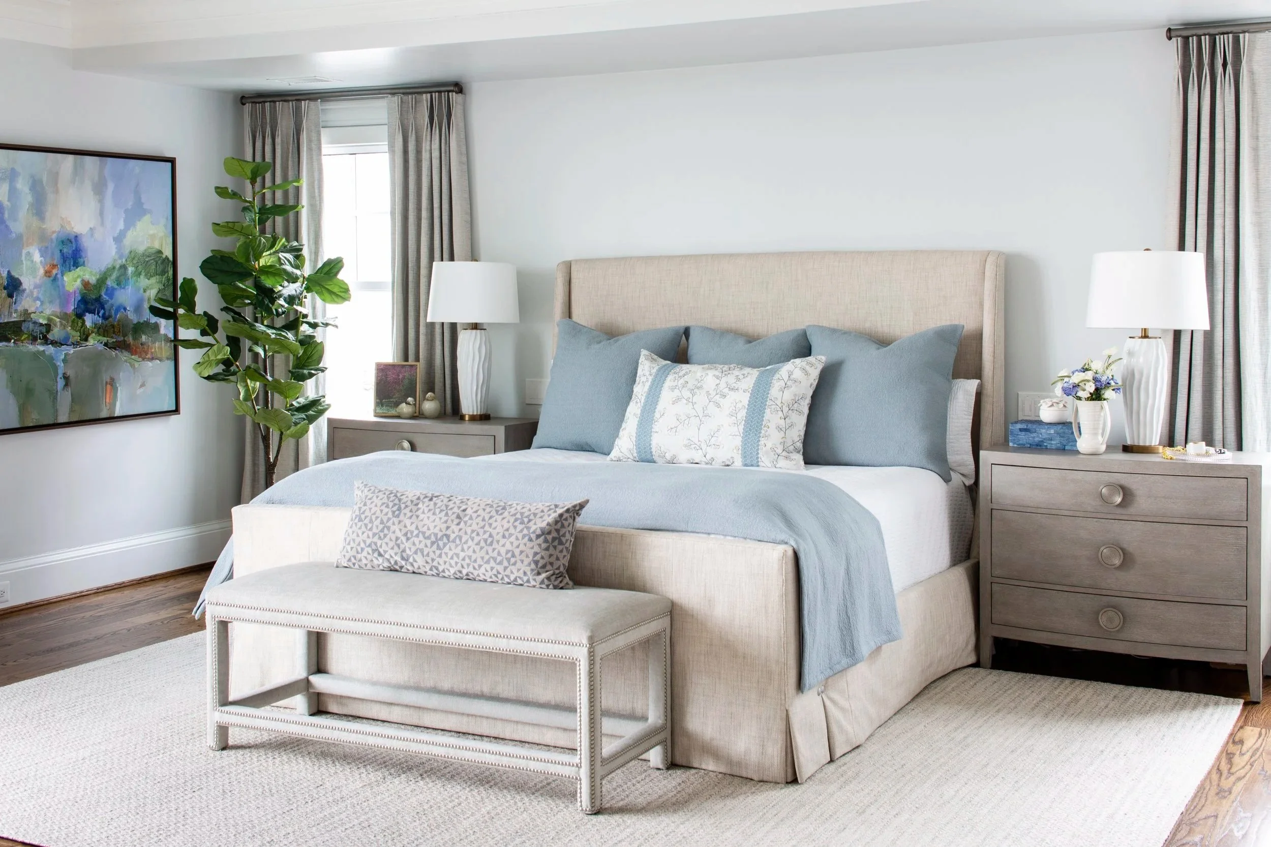 A neatly made bed with light blue and white bedding, decorative pillows, and a beige upholstered headboard in a bright bedroom with artwork, lamps, and a potted plant.