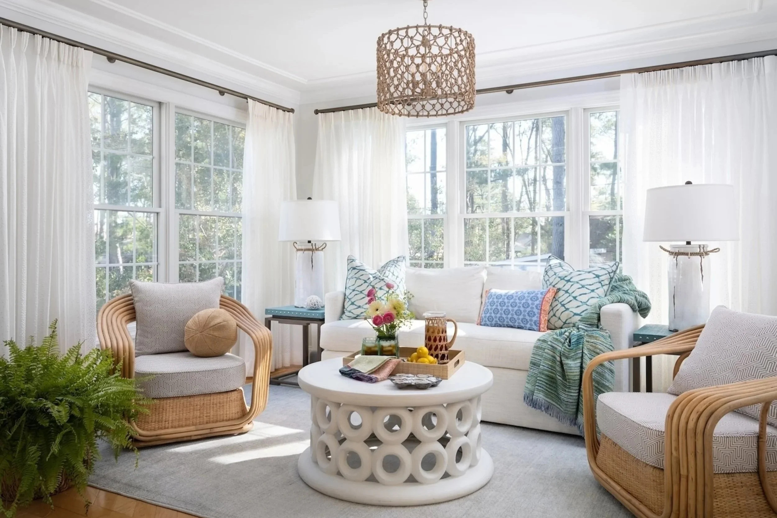 Bright living room with white sofa, rattan armchairs, large windows with white curtains, a round white coffee table, and decorative pillows.