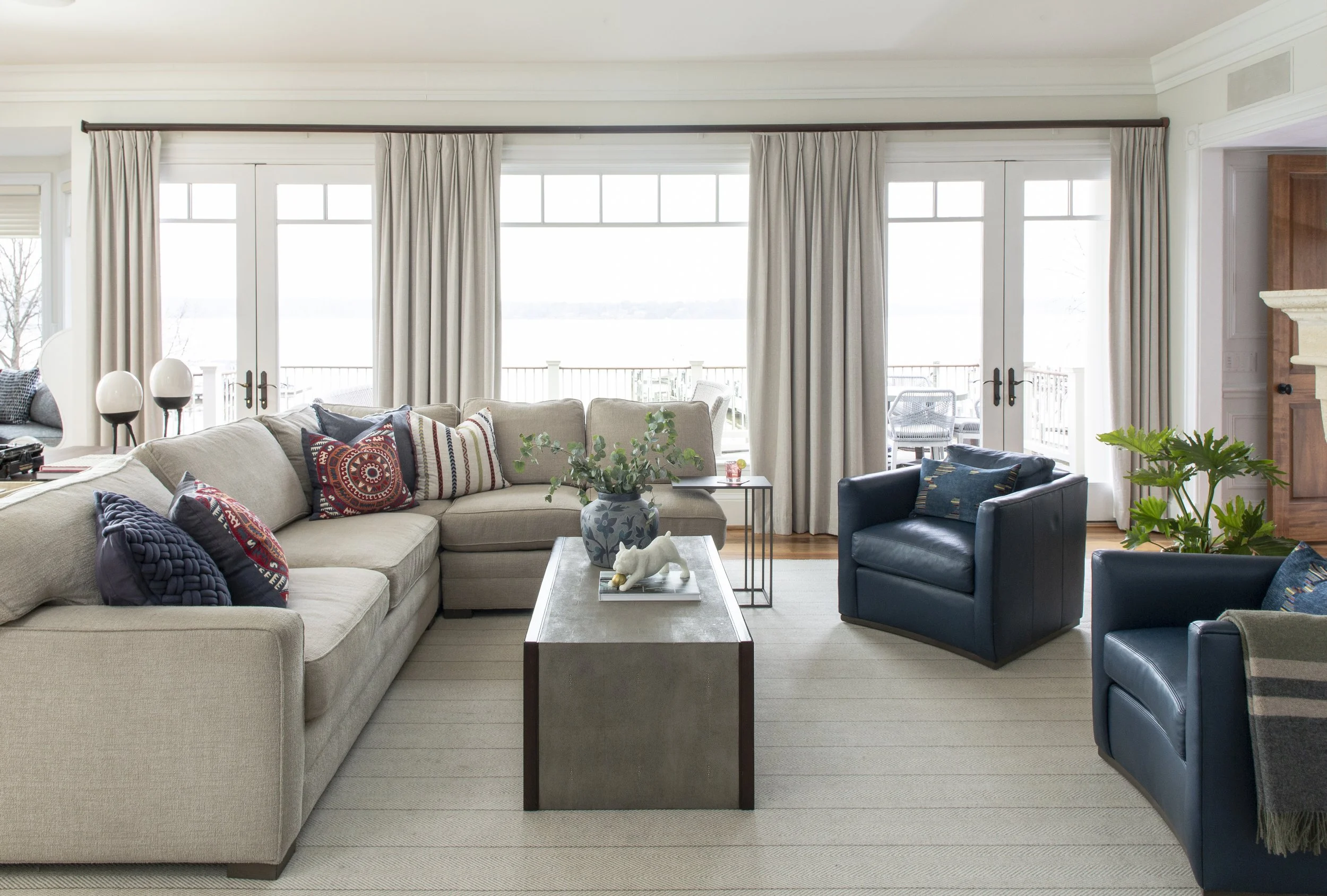 Living room with large beige sectional sofa, blue armchairs, and a wooden coffee table. There are decorative pillows, a potted plant, and a view of a balcony with an ocean in the background.