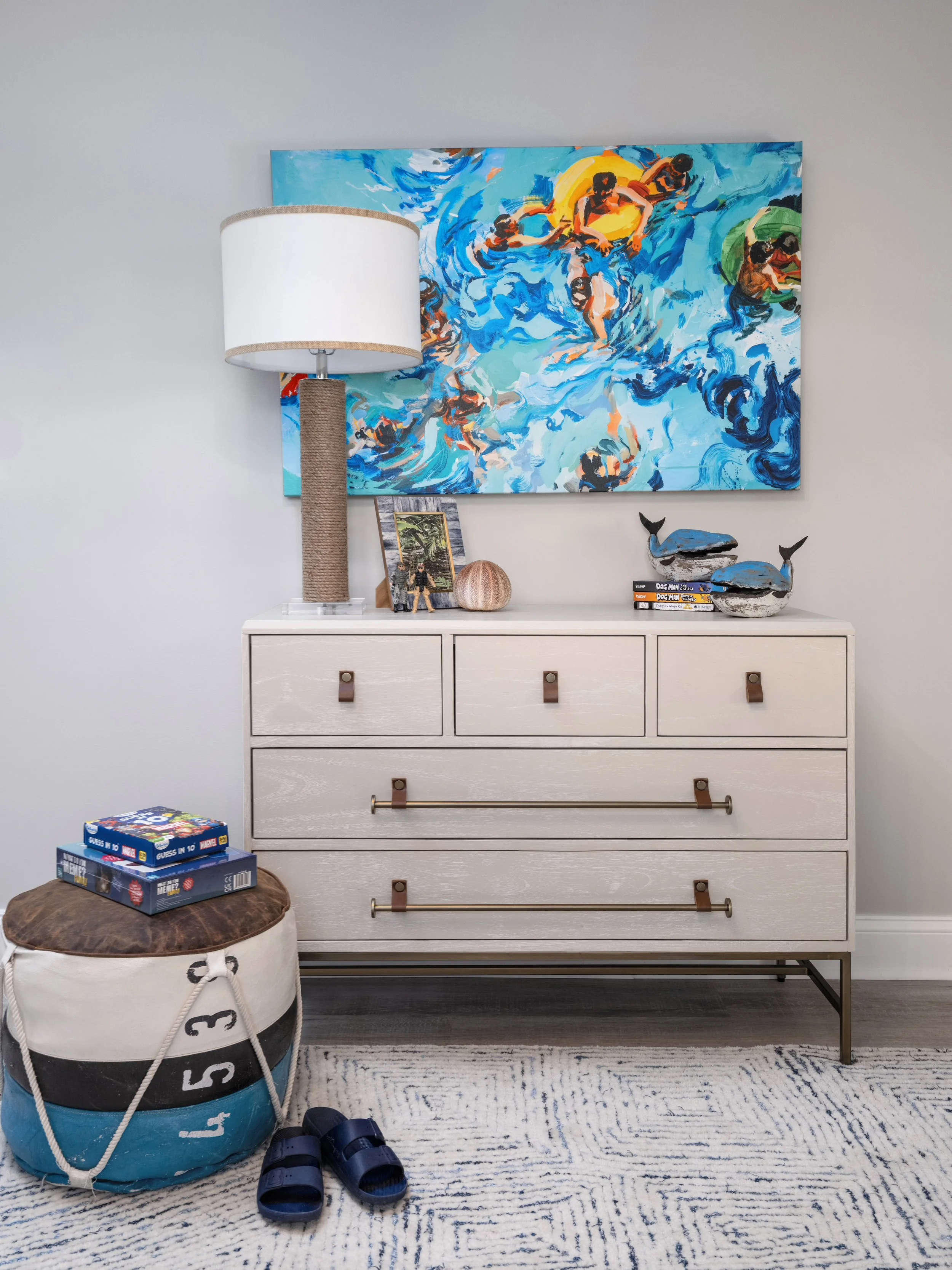 A white dresser with leather handles, a table lamp, small framed photo, decorative globe, and whale figurines on top. A colorful abstract wall art depicting children swimming is above the dresser. On the floor, there is a bean bag with numbers and a board game, alongside a pair of sandals.