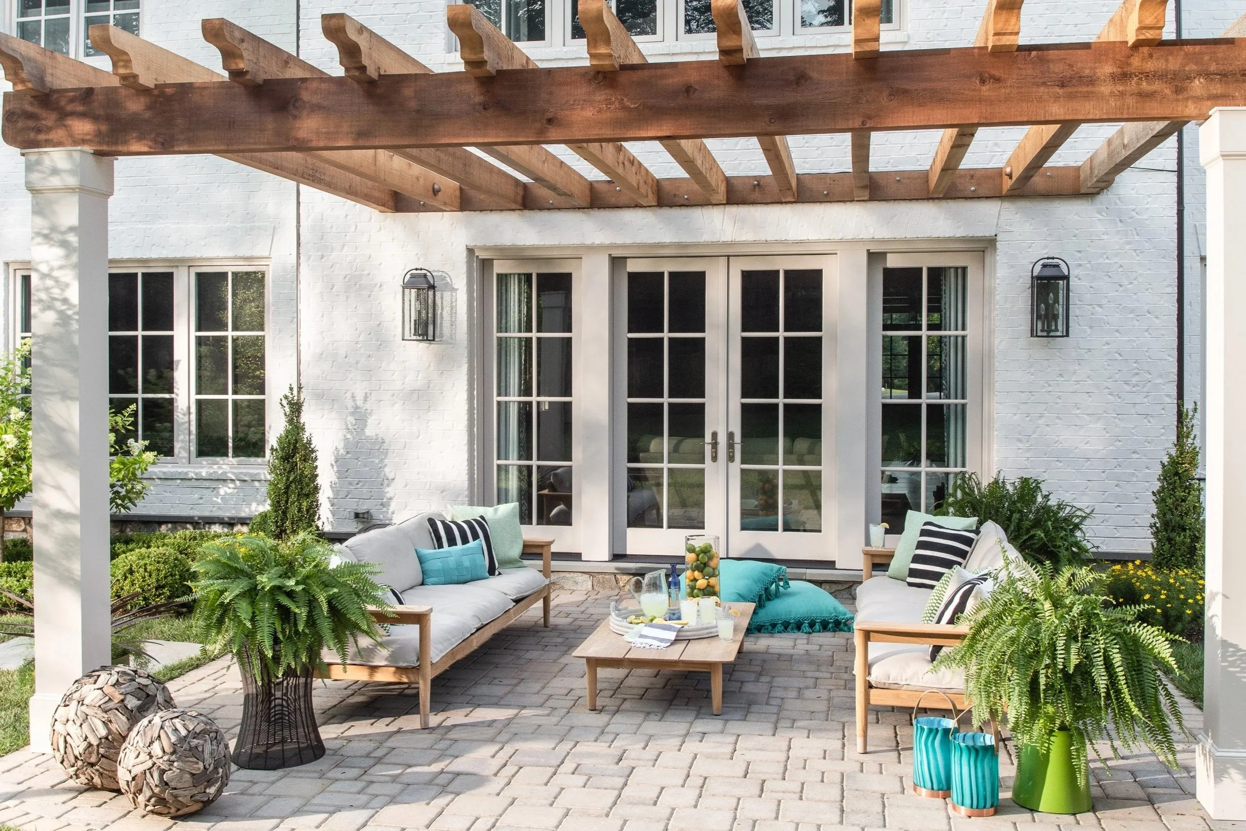 Backyard patio area with two wooden sofas, a center table, potted ferns, decorative pillows, and outdoor lighting attached to the white brick house