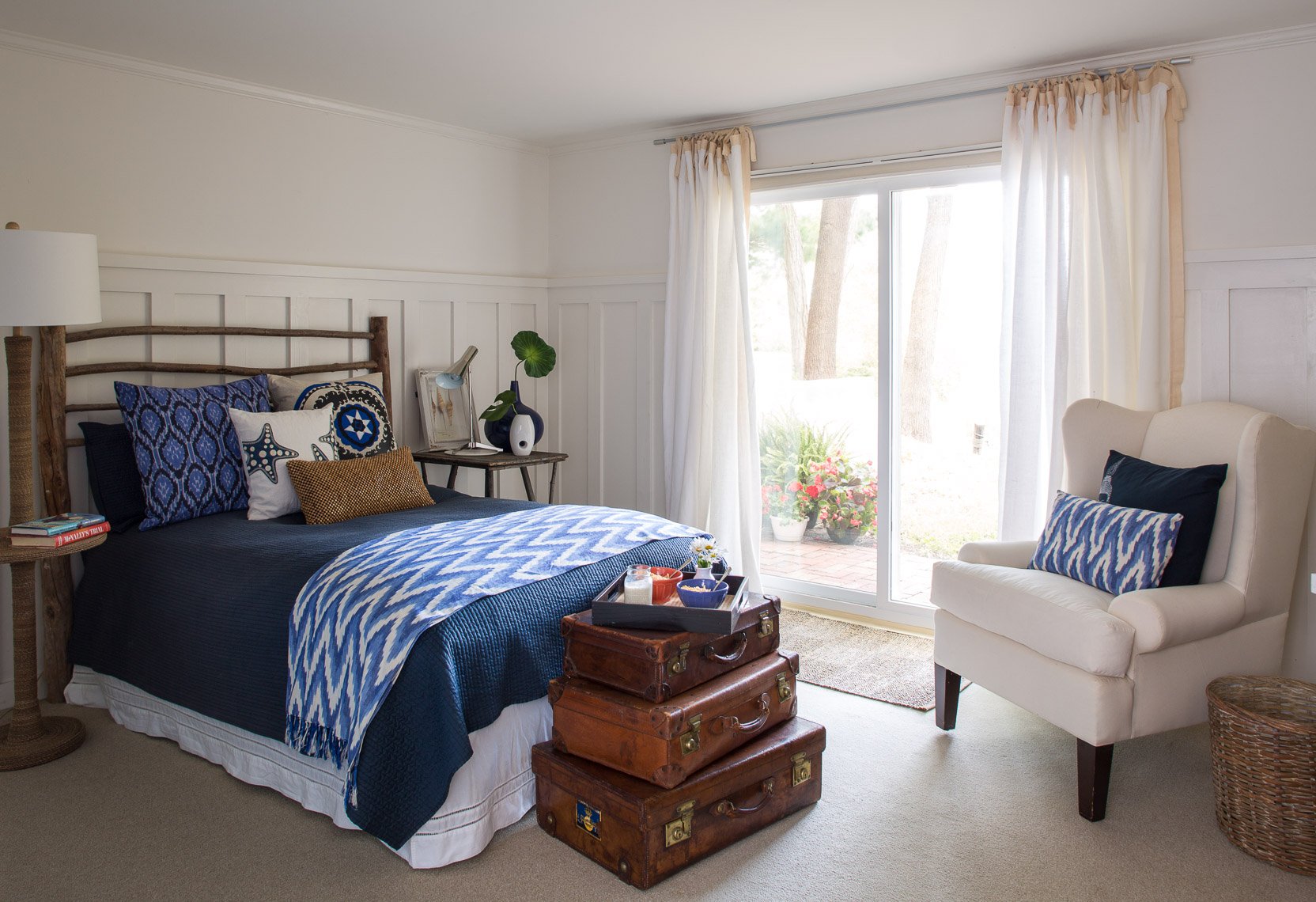 A bedroom with a bed covered in a blue quilt and decorative pillows, a white armchair with pillows, a view of a patio through sliding glass doors, and vintage suitcases serving as a coffee table.