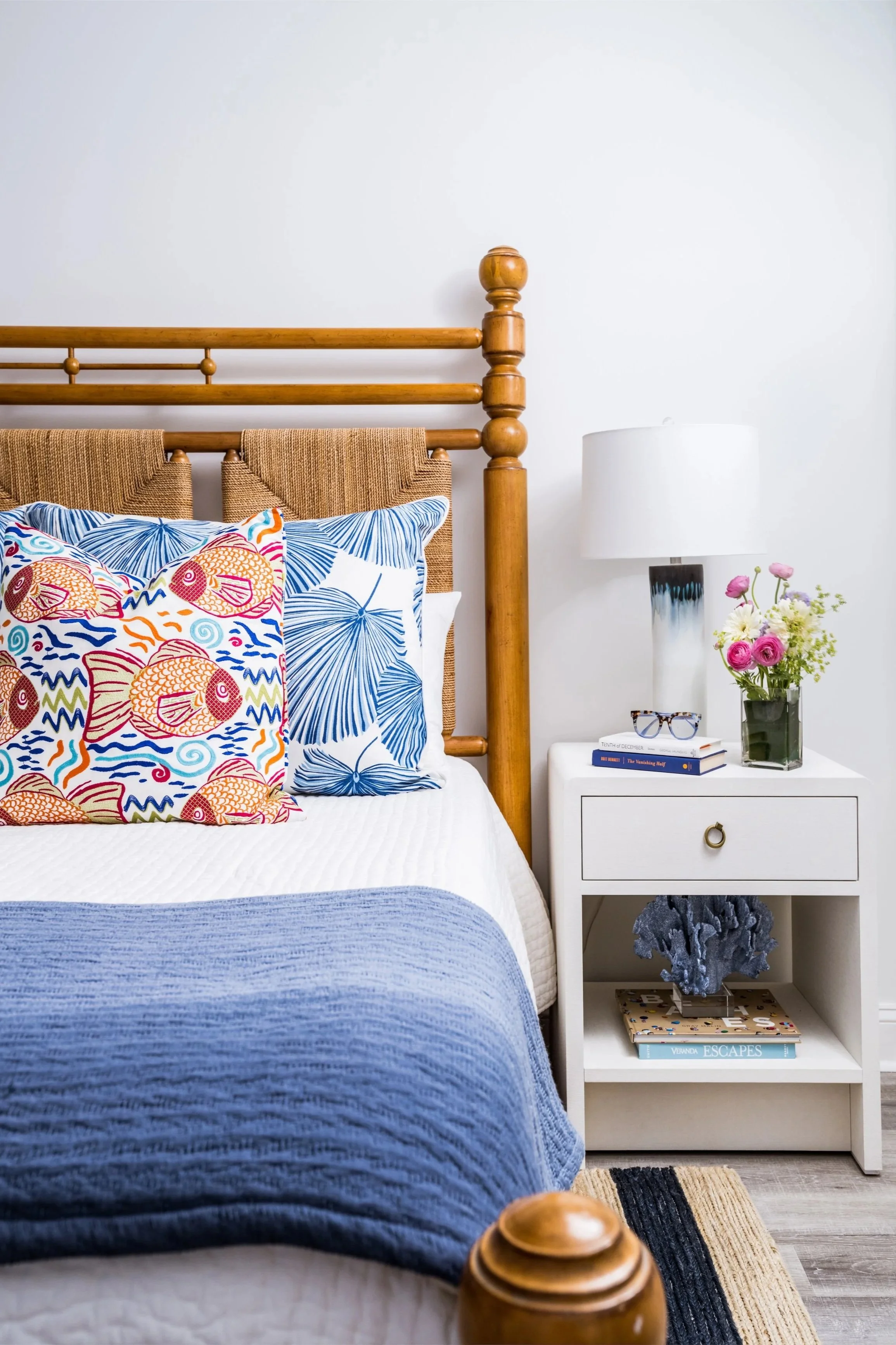 A cozy bedroom corner with a wooden bed, colorful fish-themed and blue leaf-patterned pillows, a white nightstand with a vase of pink and white flowers, books, a gray coral sculpture, and a white table lamp.