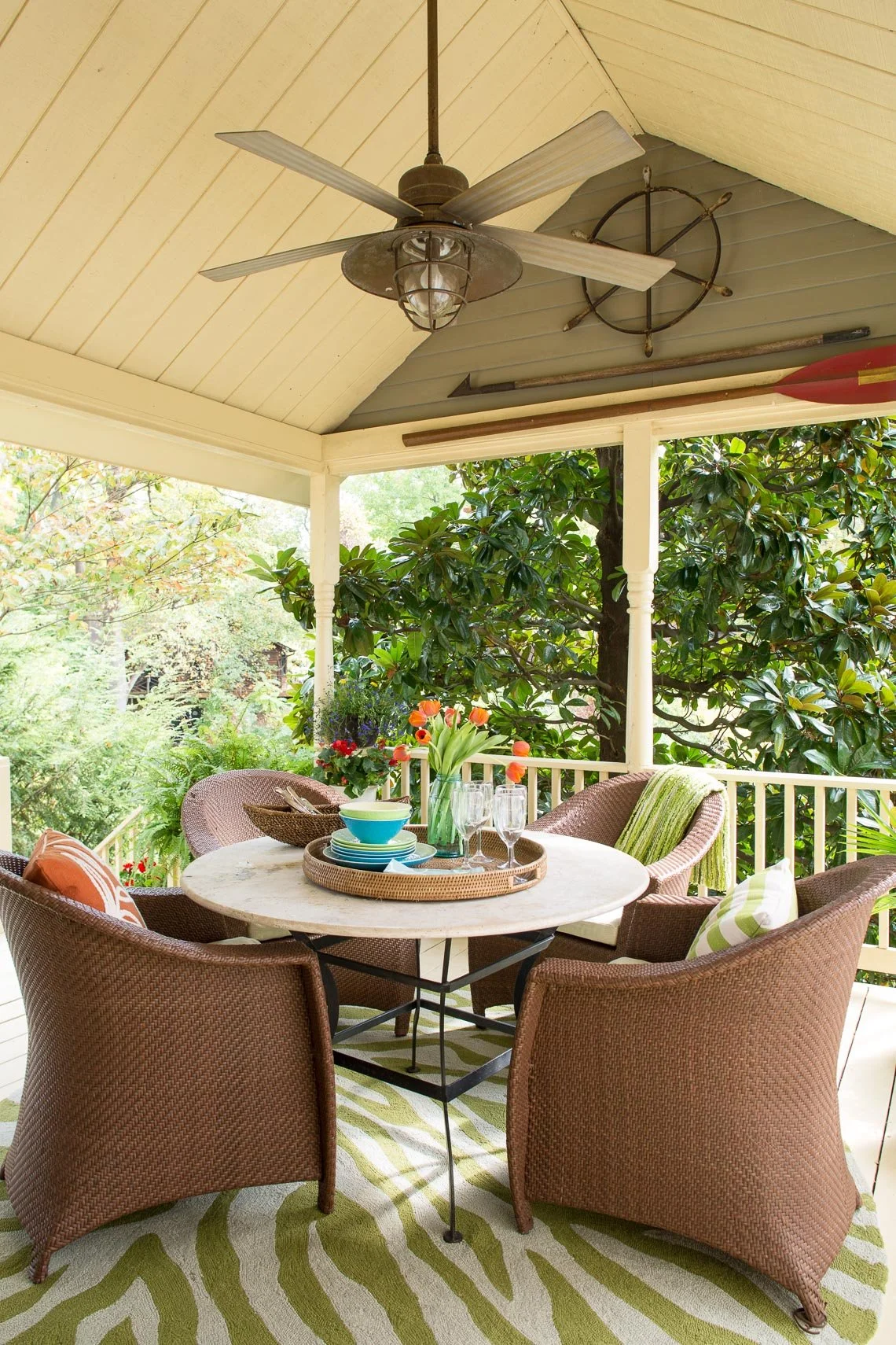 Outdoor covered patio with a round table set with bowls, glasses, and a vase of tulips, surrounded by wicker chairs, with a ceiling fan and nautical decorations overhead, and greenery in the background.