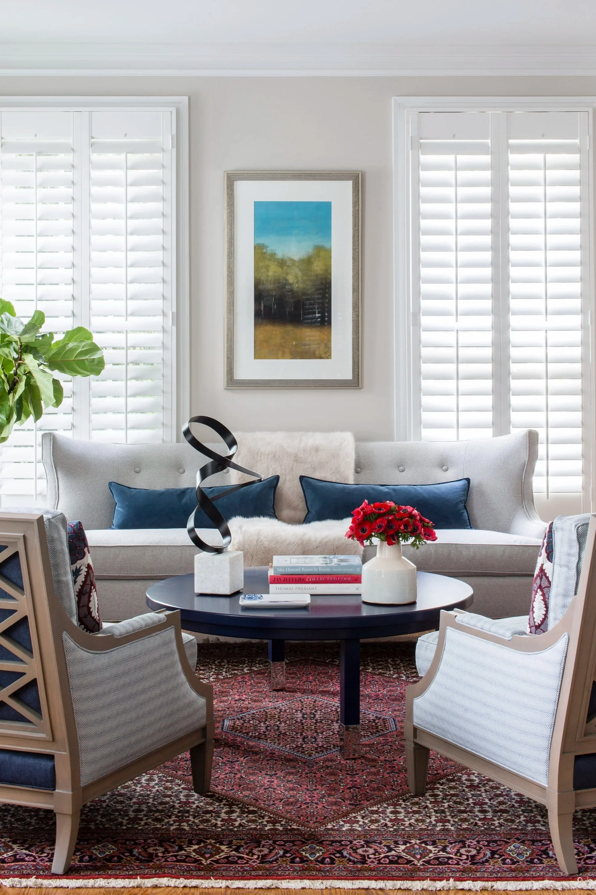 Living room with white walls, large windows with white shutters, a white upholstered sofa with blue pillows, a round black coffee table with decorative items including a black abstract sculpture, a white vase with red flowers, and books. There are two light-colored armchairs with patterned cushions and an ornate red and black area rug. Framed landscape painting on the wall and a large green leafy plant on the side.