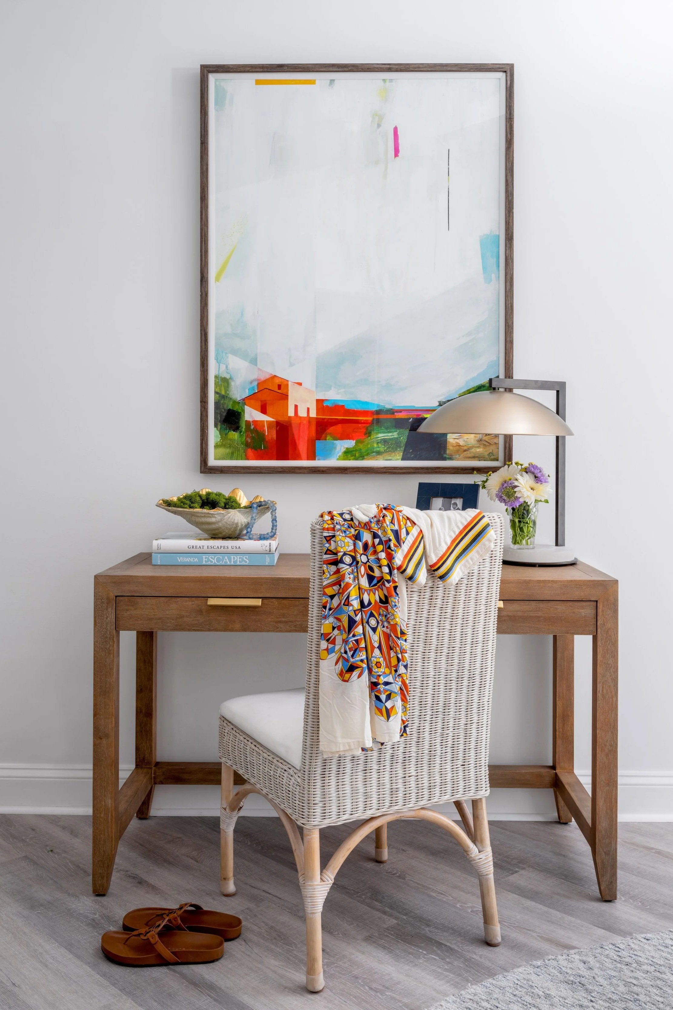 A cozy workspace with a wooden desk, rattan chair, colorful textile, and modern lamp, featuring a large colorful abstract painting on the wall.