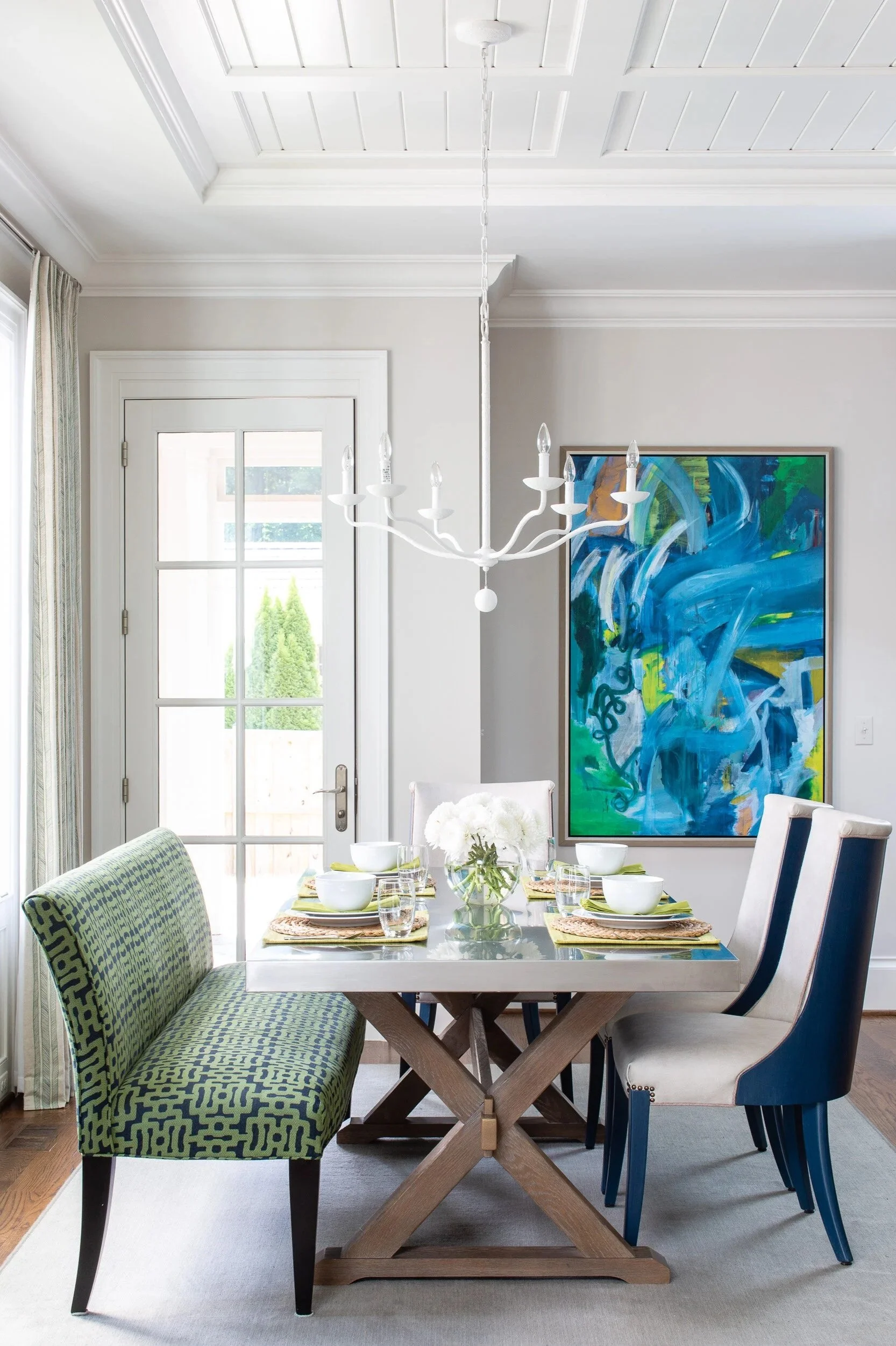 A dining room with a white table set with four bowls, four glasses, and a vase of white flowers, surrounded by a green patterned bench and white and blue upholstered chairs. A white chandelier hangs above, and an abstract blue painting is on the wall. A door with glass panels leads outside, and sunlight streams in.