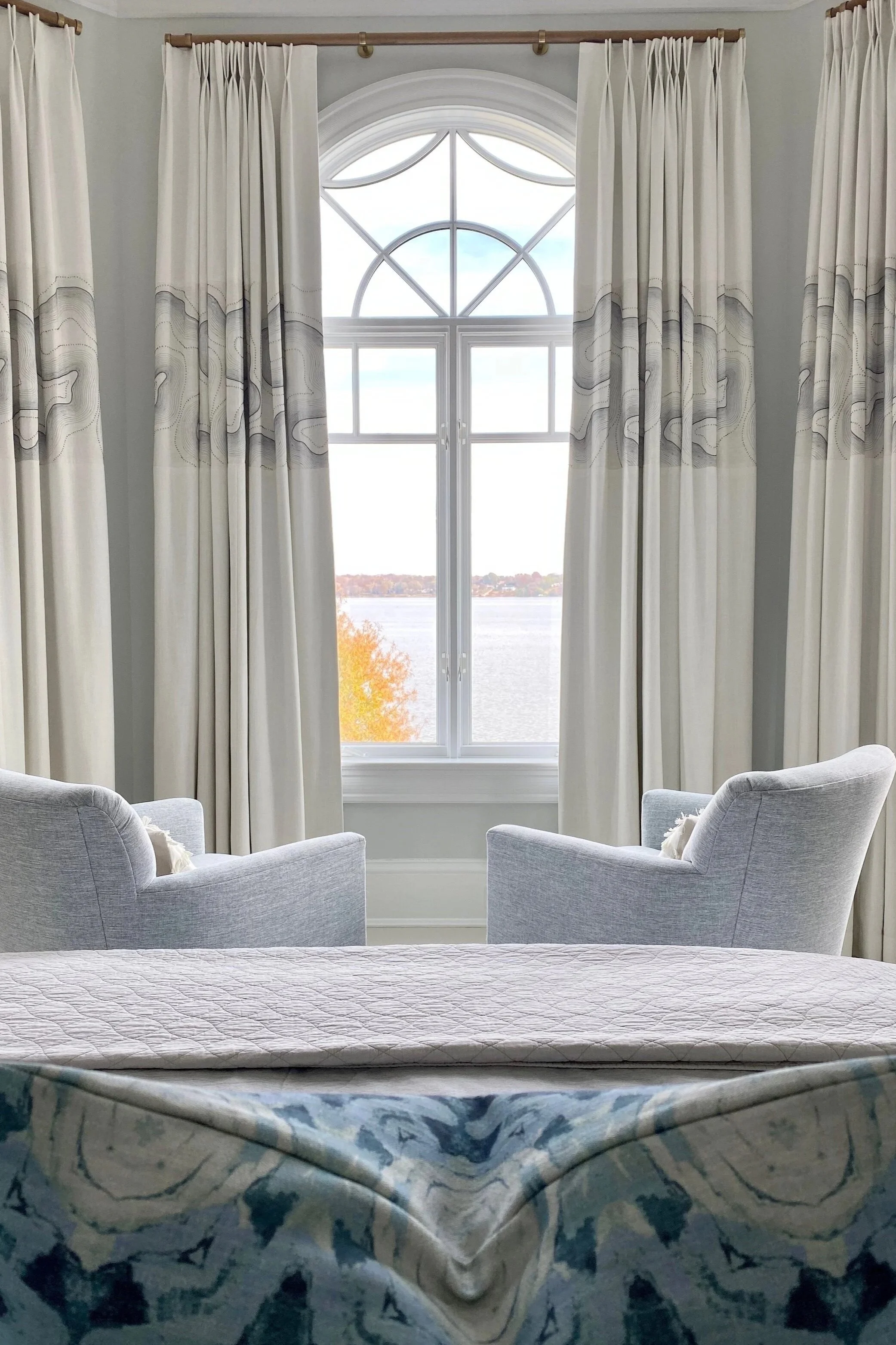 A room with a large arch window showing a view of a lake and autumn trees. There are two light gray armchairs facing the window, with white curtains on either side.