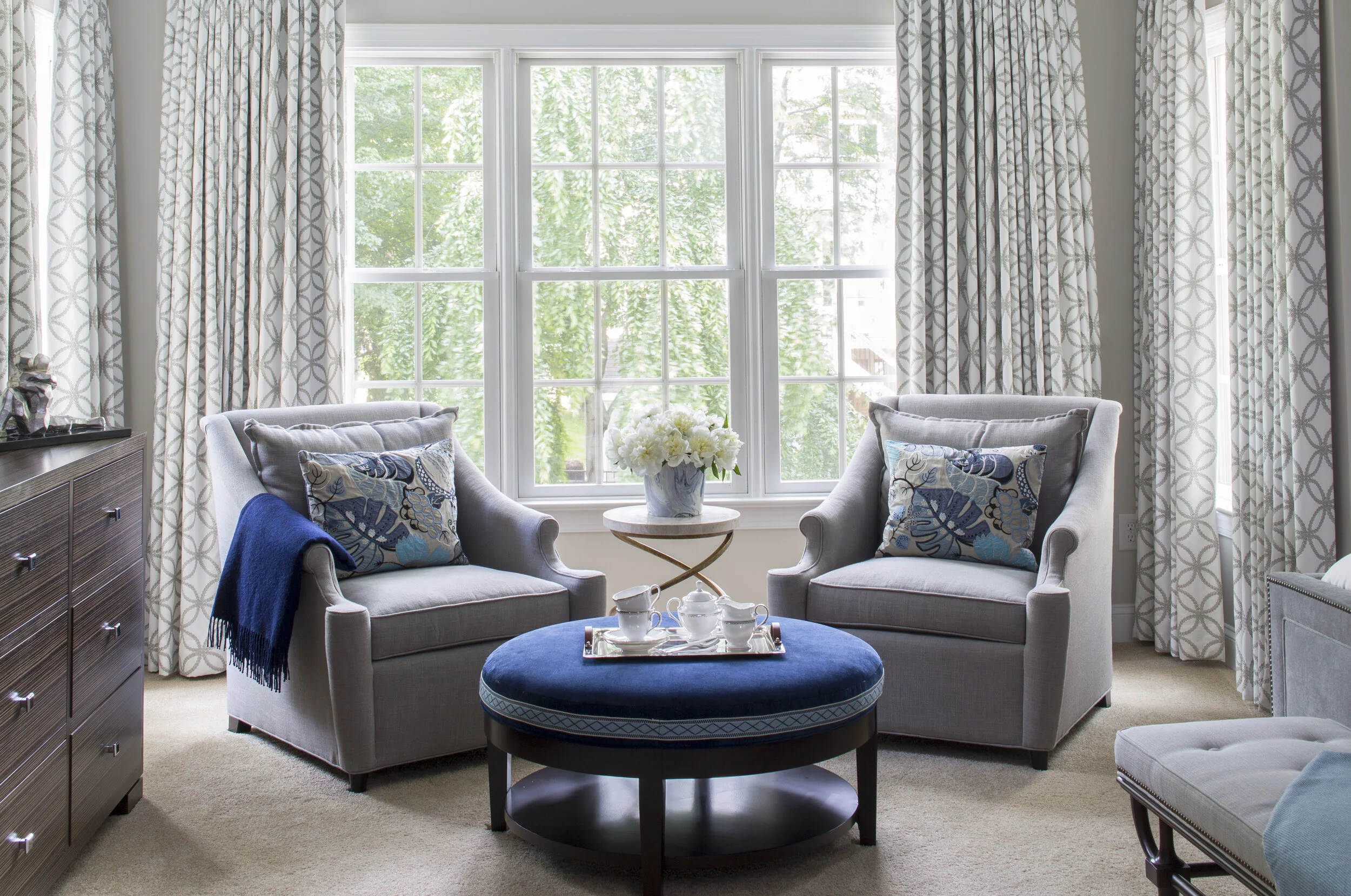 A bright living room with large window, light-colored armchairs with patterned pillows, a round blue ottoman with tea set, a side table with white flowers, and a dresser, with curtains featuring a geometric pattern.