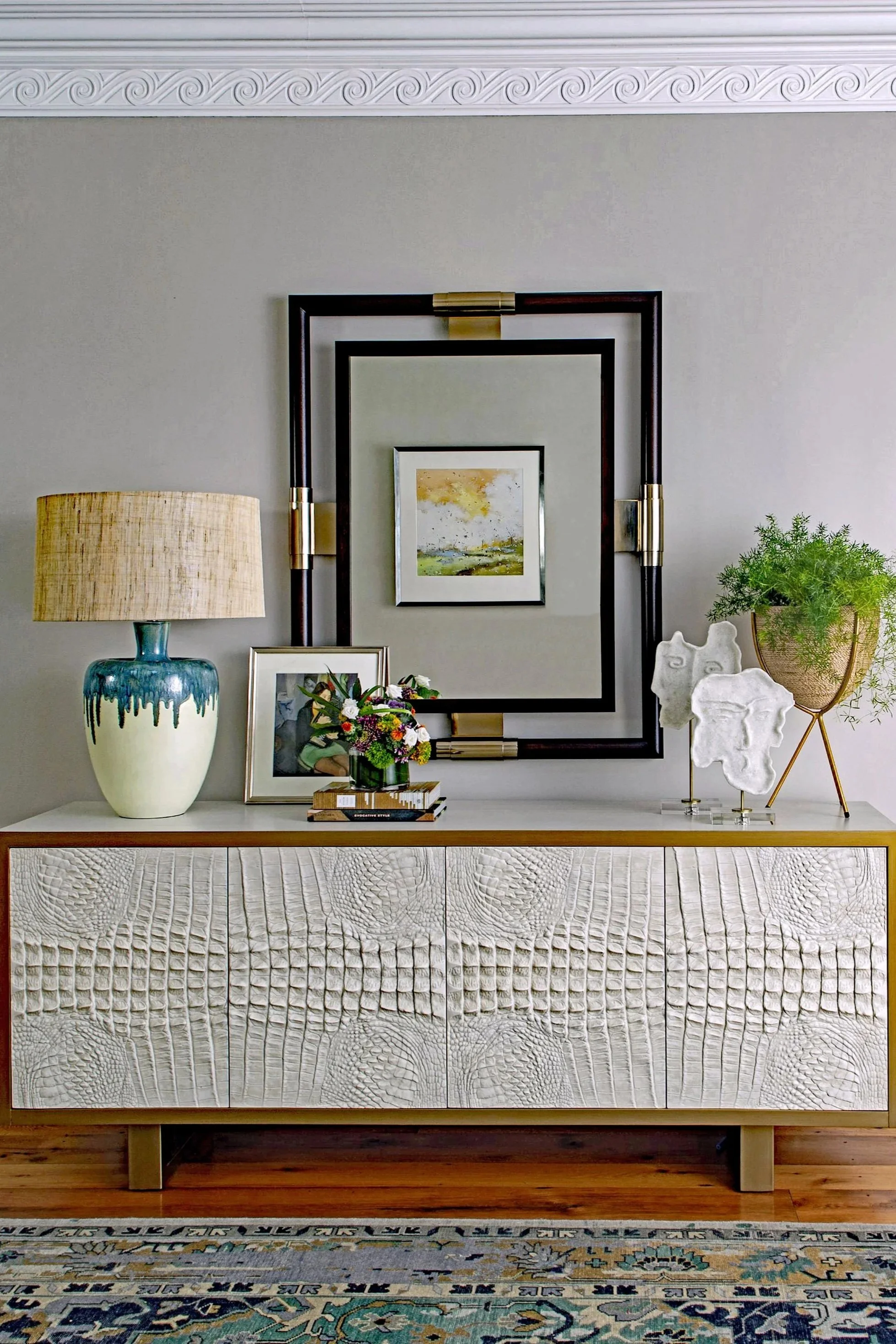 A beige sideboard with textured, pressed patterns on the front, sitting on wooden legs, decorated with a large ceramic lamp with a beige shade, a framed photograph, a bouquet of flowers, a stacked set of books, and a tall potted plant with green foliage, with a large geometric mirror and small abstract sculptures on top.