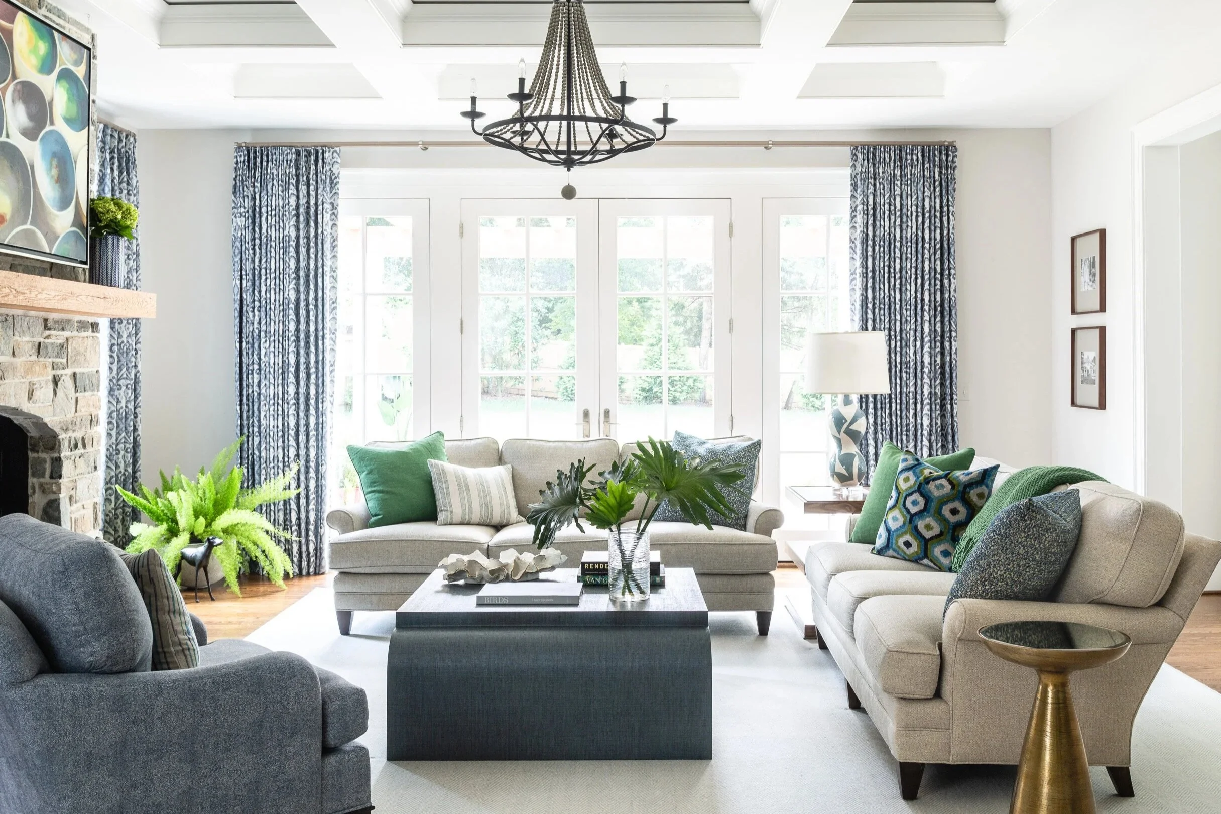 Bright living room with two beige sofas and a gray armchair, coffee table with books and plants, large windows with blue patterned curtains, a chandelier, and decorative accents.