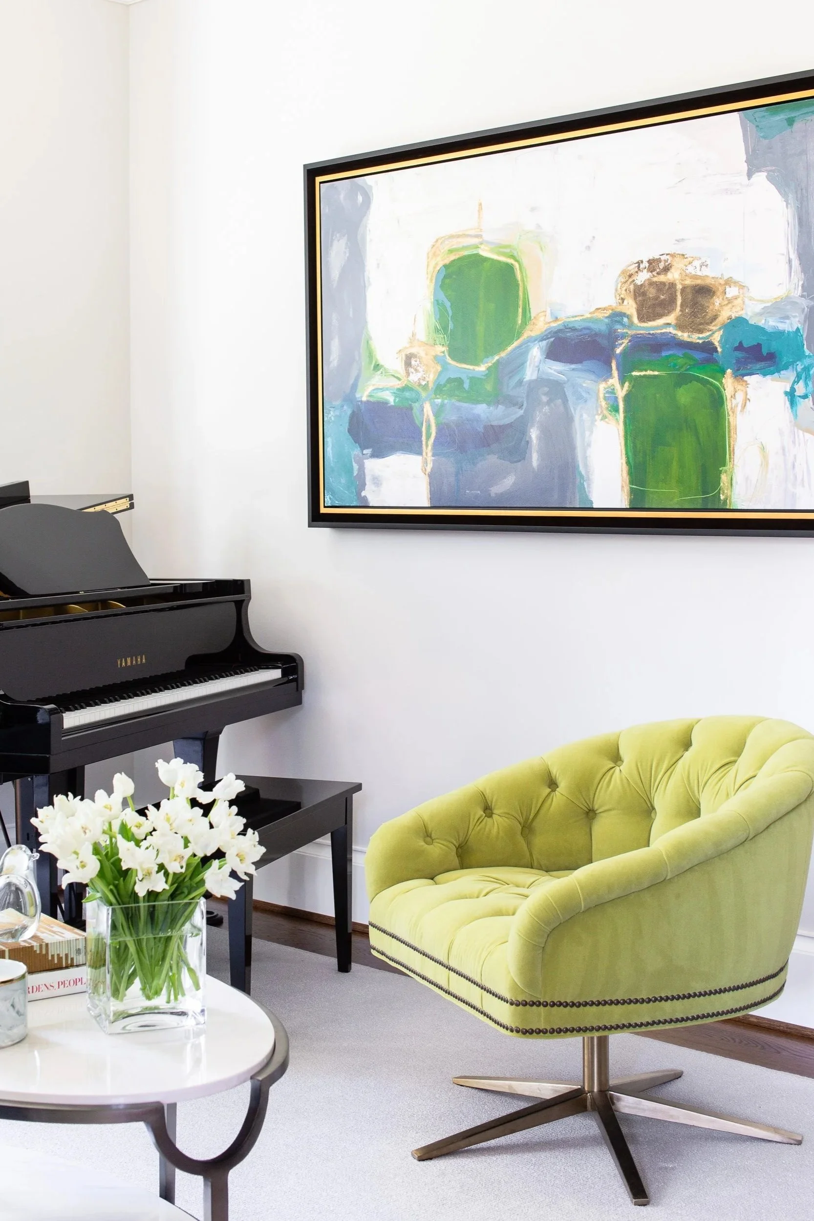 Interior of a room with a black Yamaha piano, a white table with a glass vase of white flowers, a bright yellow-green upholstered swivel chair, a large abstract painting on the wall with green, blue, and beige colors.