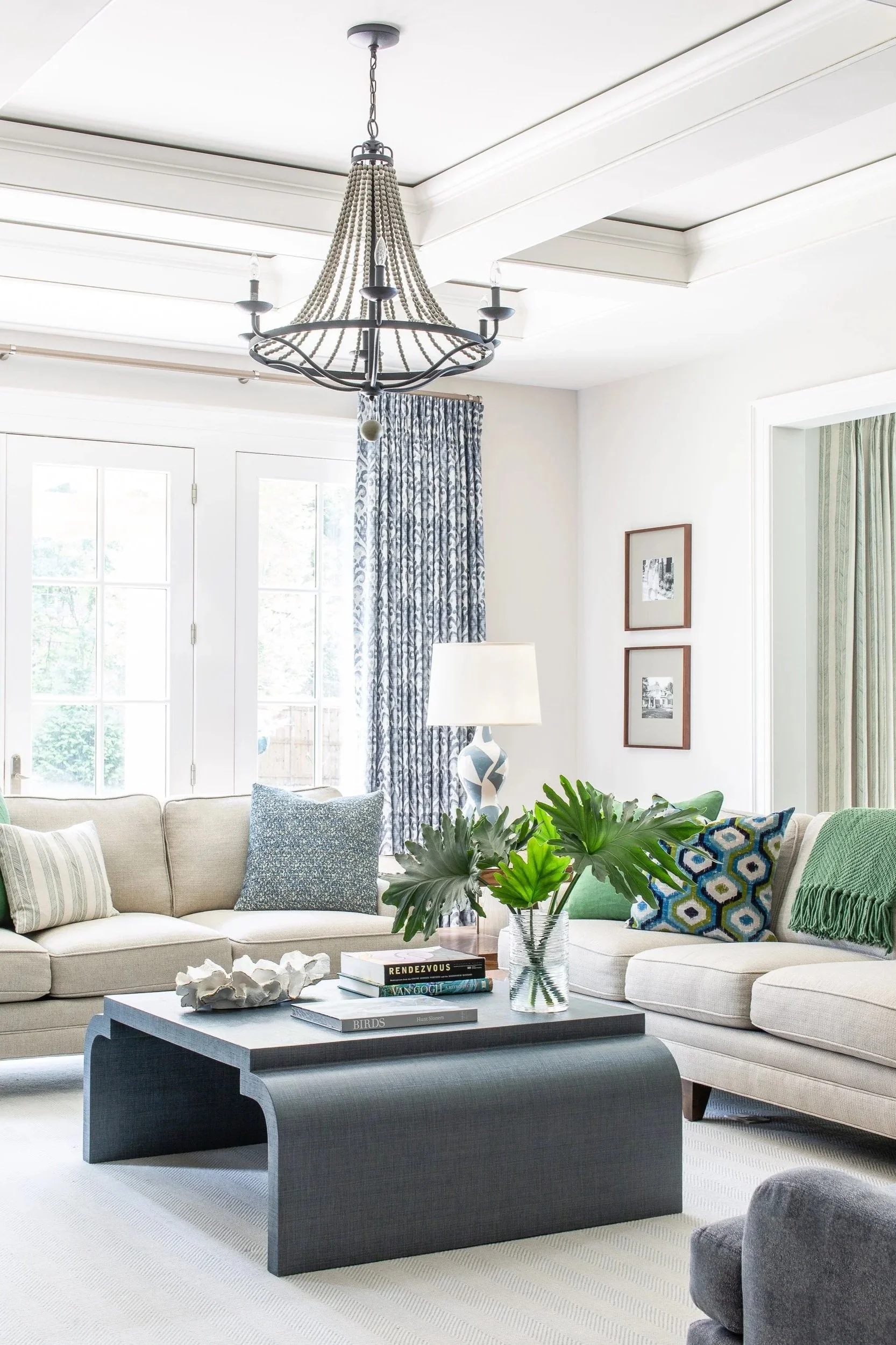 A bright living room with a beige L-shaped sofa adorned with patterned throw pillows, a black contemporary coffee table with a vase of large green leaves, a table lamp with a decorative base, and patterned curtains. A chandelier hangs from the ceiling.