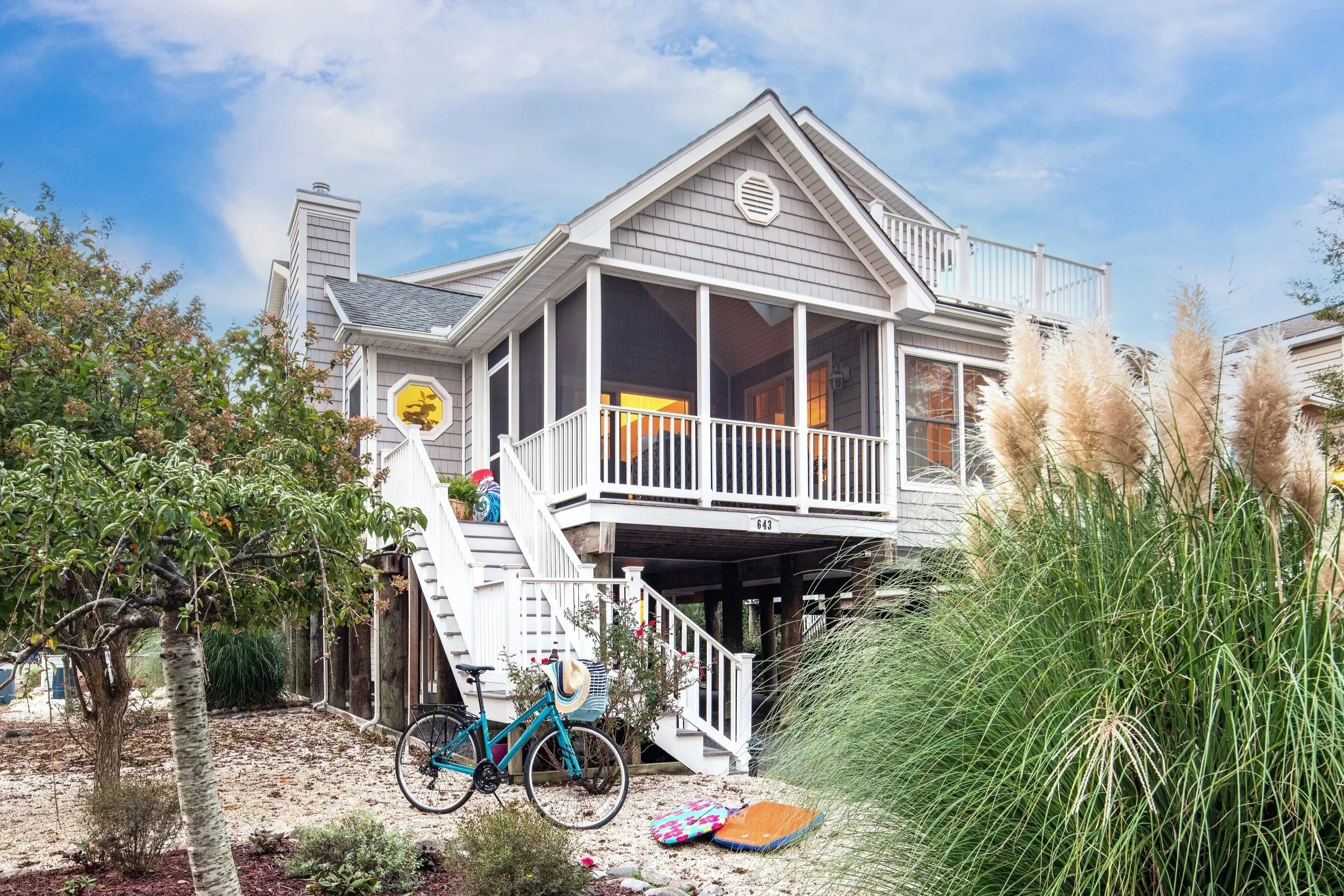 A raised beach house with a screened porch, gray siding, white railings, and a gabled roof. The house has a staircase leading up to the porch, a bicycle parked at the base, and a yard with trees, tall grass, and various outdoor items.