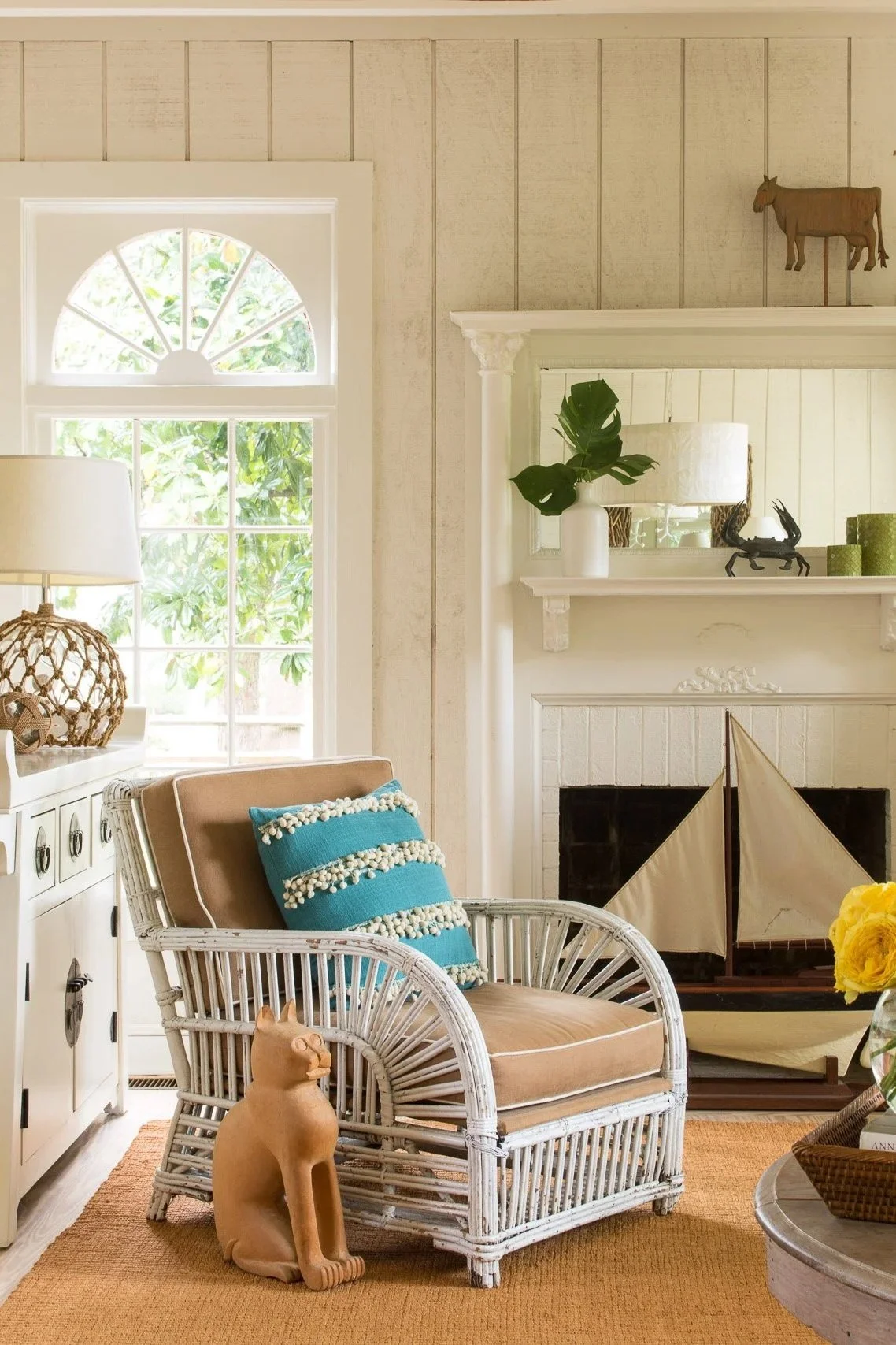 Cozy living room with a wicker armchair, a beige cushion, and a blue pillow with pom-pom trim. A tan cat-shaped sculpture sits on the floor next to the chair. There is a fireplace with decorative items on the mantel and a ship model inside. A tall window with an arched top lets in natural light, and part of a white cabinet and a lamp with a woven base are visible. There are also wall-mounted wooden animal figures and a large green leaf plant in a white vase on the mantel.