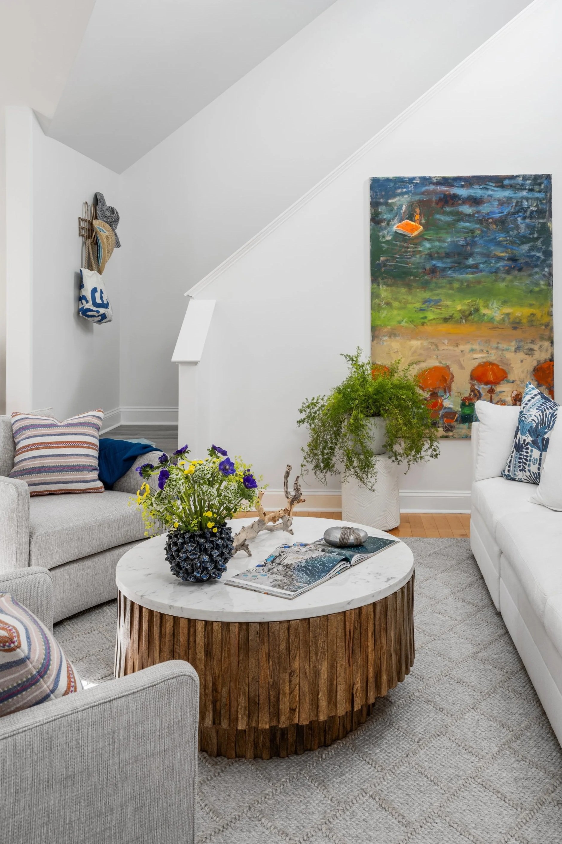 Living room with a white couch, a gray couch, and a round coffee table with a potted plant, a decorative stone, and an open magazine. Artwork hangs on the wall behind the green plant, and a coat rack with hats and a bag is on the wall to the left.