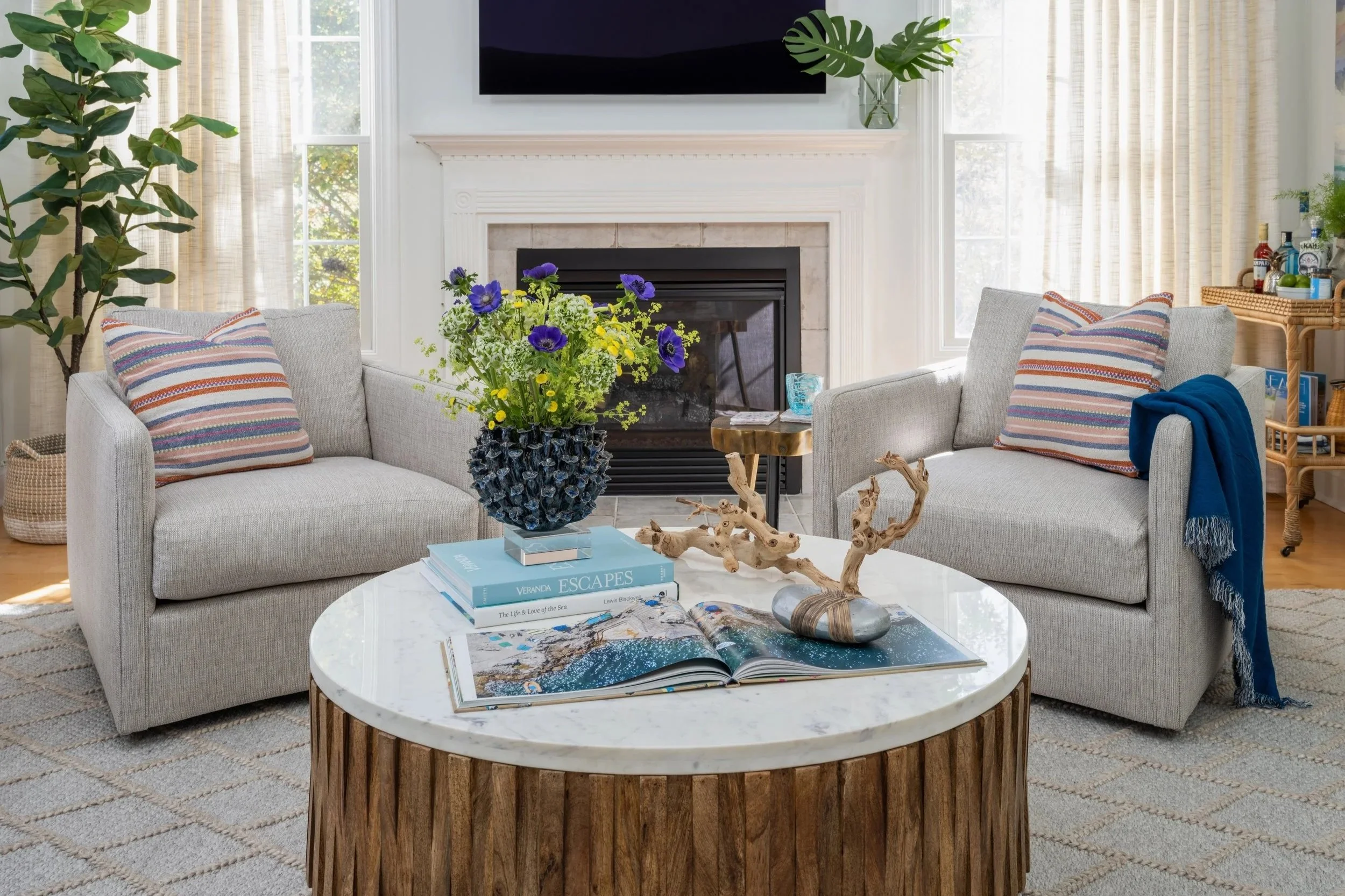 Living room with two light gray sofas with colorful striped pillows, a white marble coffee table with decorative items and books, a fireplace with a television mounted above, large windows with beige curtains, and various plants and decor.