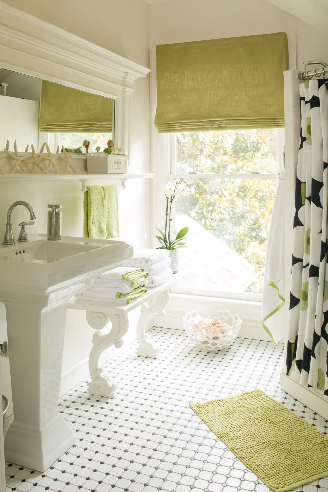 Bright bathroom with white hexagon tile floor, white sink, green window shade, and black and white shower curtain. Green towel, vase with orchid, wicker basket, and bathroom accessories are visible.