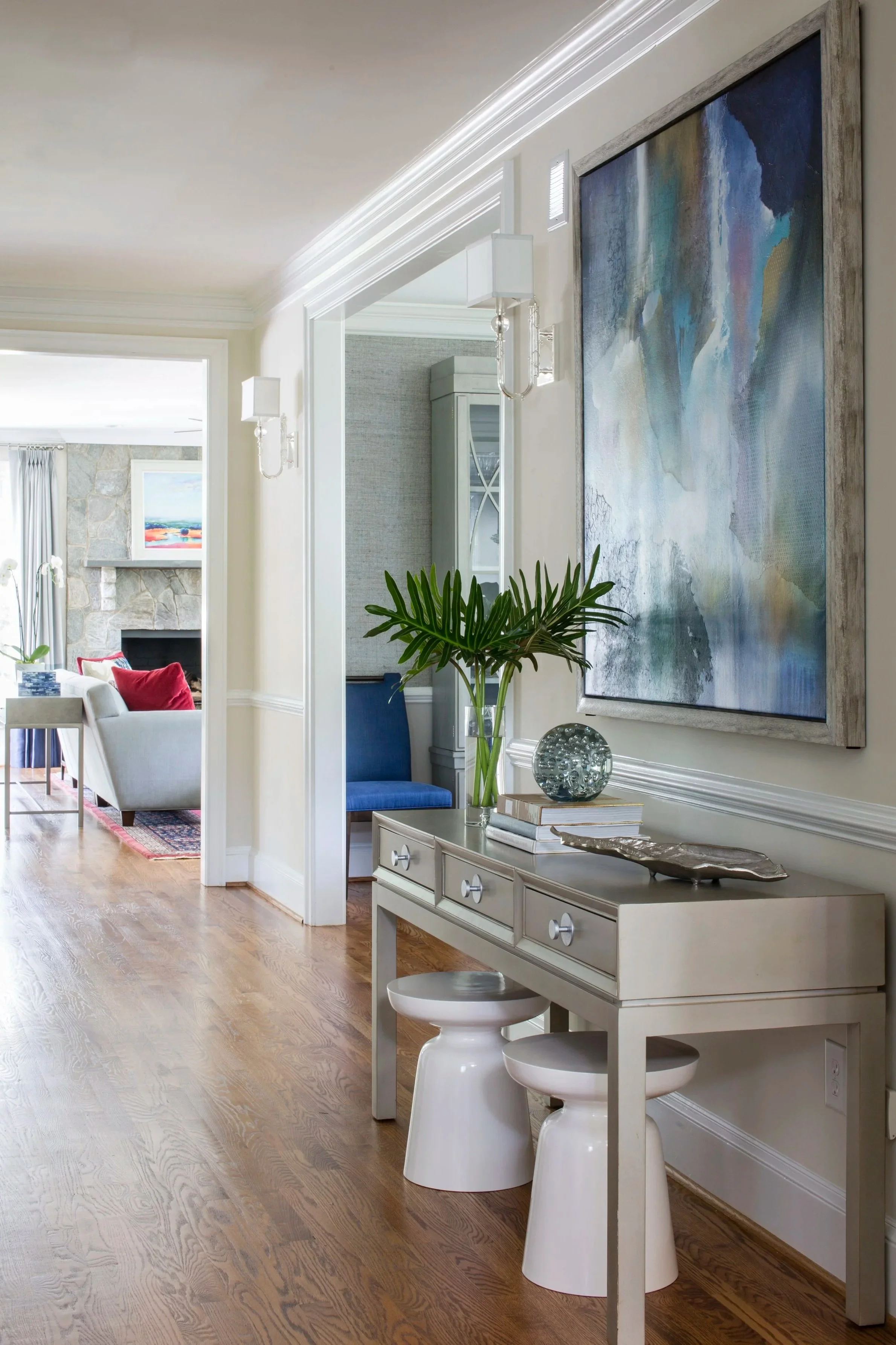 Interior view of a living space with a hallway, artwork, a console table with decorative items, and glimpses of a living room with a fireplace and blue chair.