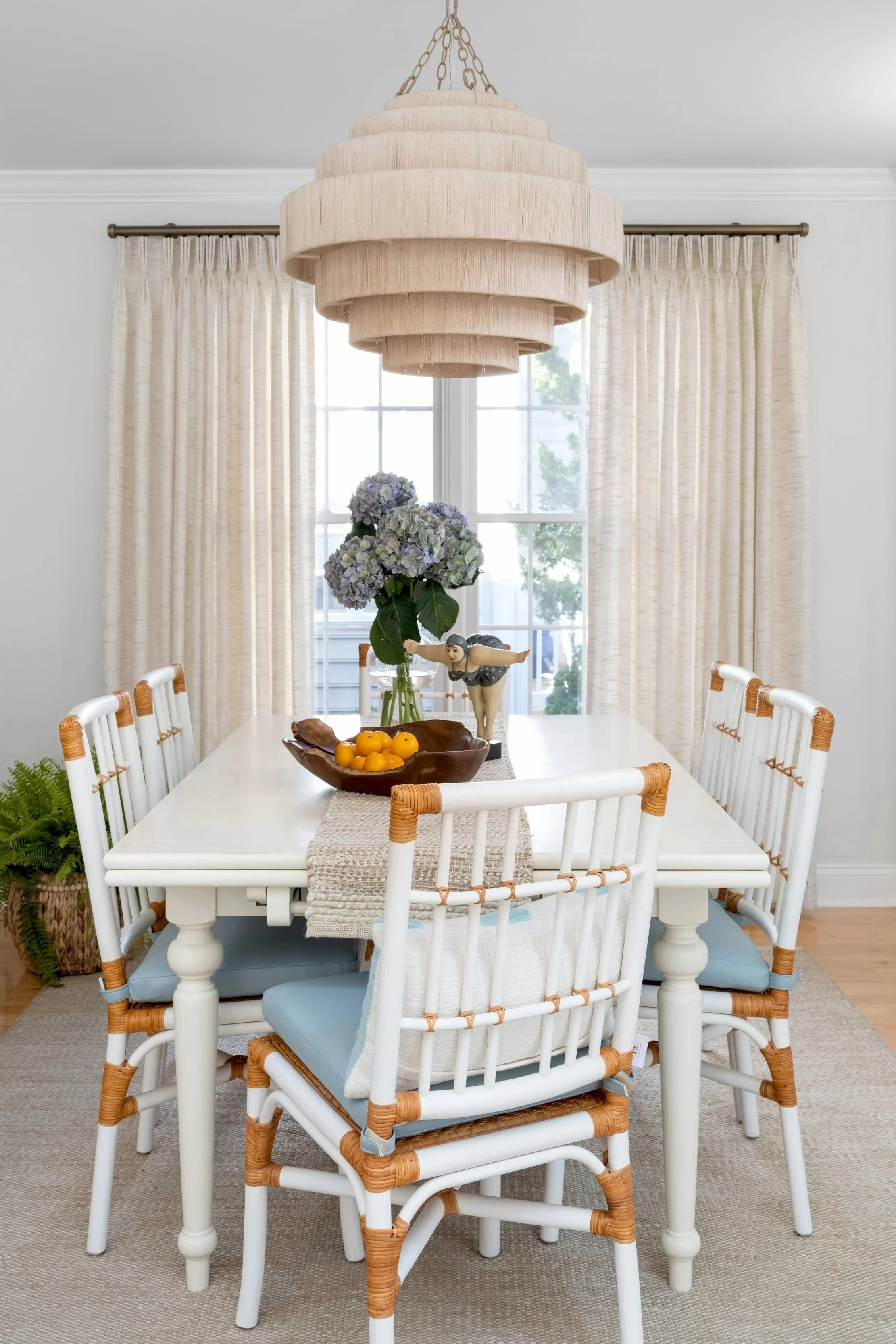 A dining room with a white table, six white chairs with blue cushions, beige curtains, a large beige pendant light fixture, a centerpiece with purple hydrangeas, a wooden bowl of oranges, a small figurine, and a pot of greenery in the corner.