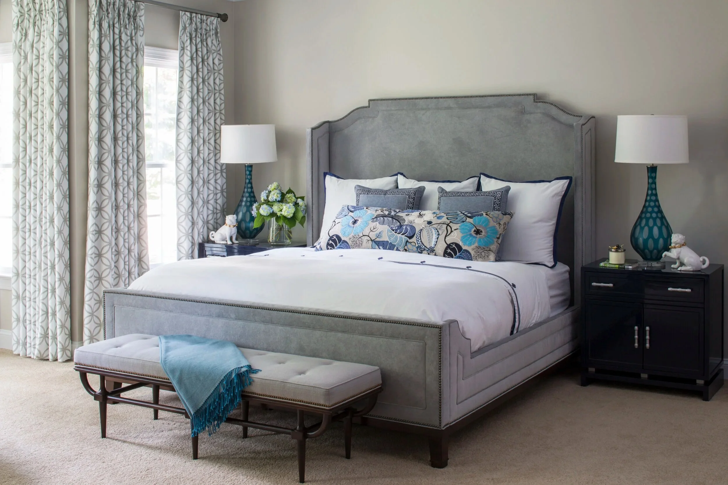 Bedroom scene with a large bed featuring a grey upholstered headboard, white bedding with blue and grey pillows, two black nightstands with matching blue lamps, a bench at the foot of the bed with a blue throw blanket, and floor-to-ceiling patterned curtains next to windows.