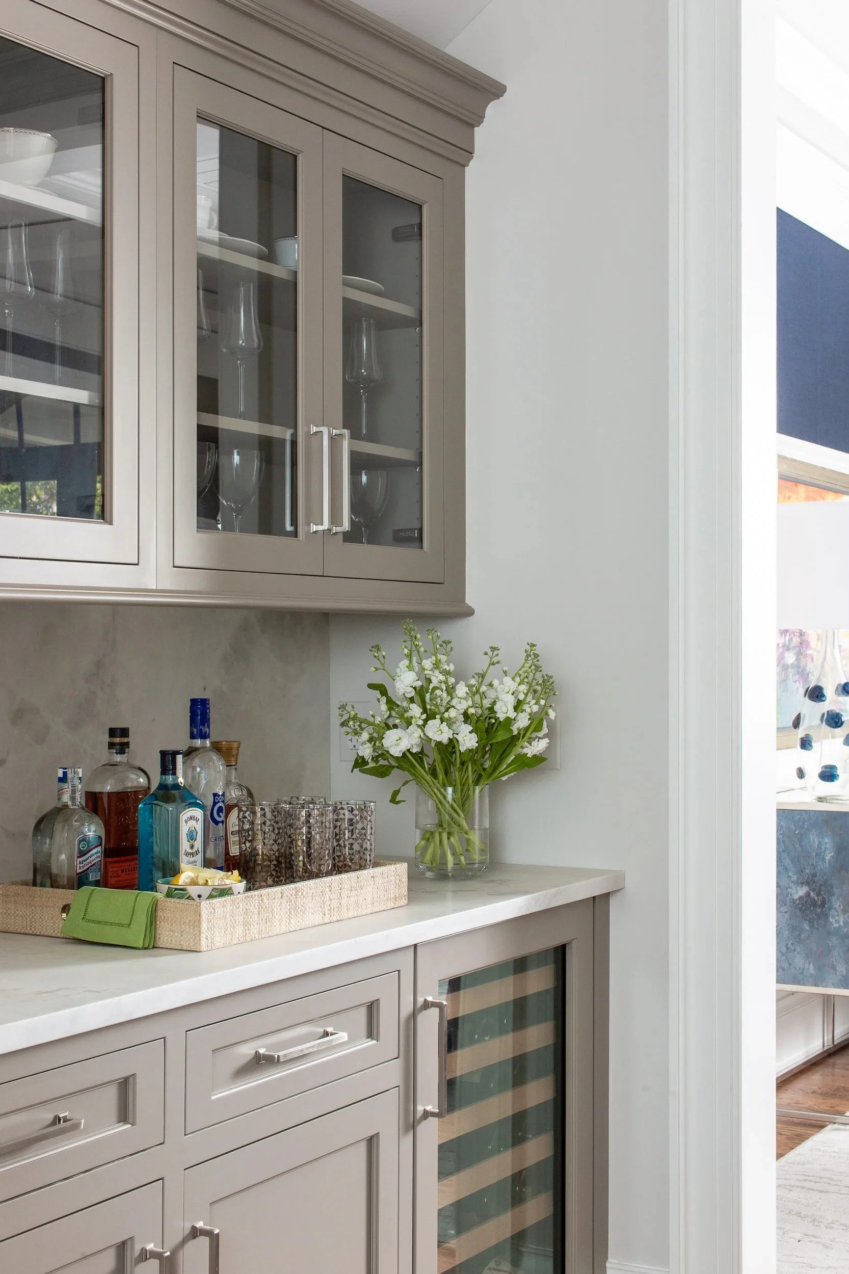 A kitchen counter with a bouquet of white flowers in a glass vase, several liquor bottles, and drinking glasses, with beige cabinets above and below, and a sliding glass door to the right.