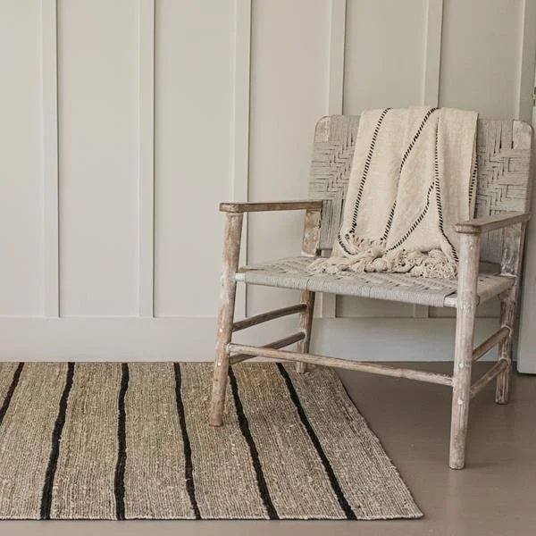 Rustic wooden chair with a woven seat and back, draped with a fringed cream blanket, beside a striped woven rug, set against a paneled wall.